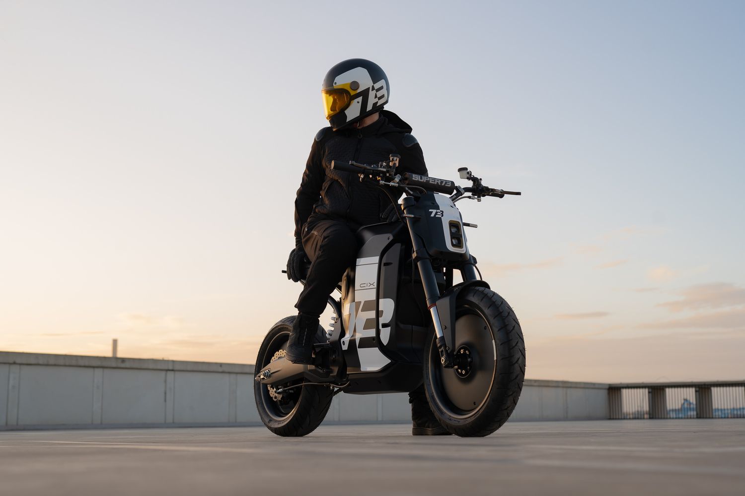 Motorcycle rider in black gear performs stunts on a sleek custom bike against an urban backdrop at sunset.