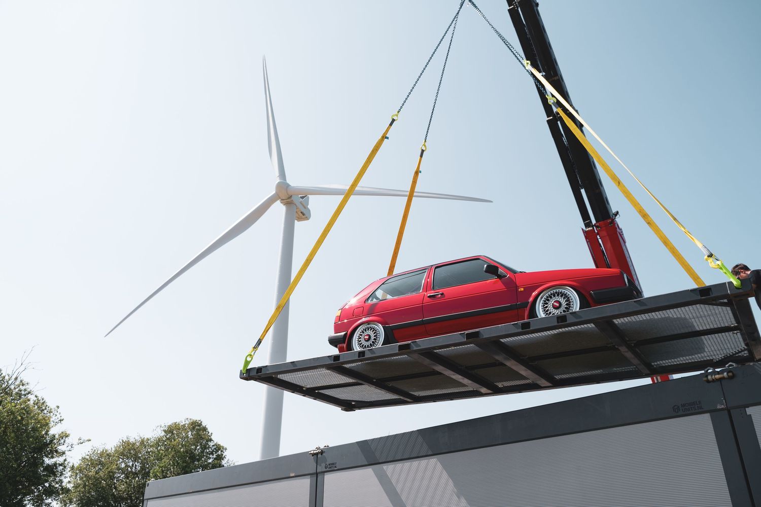 Red vintage car being lifted by crane with wind turbine in background.