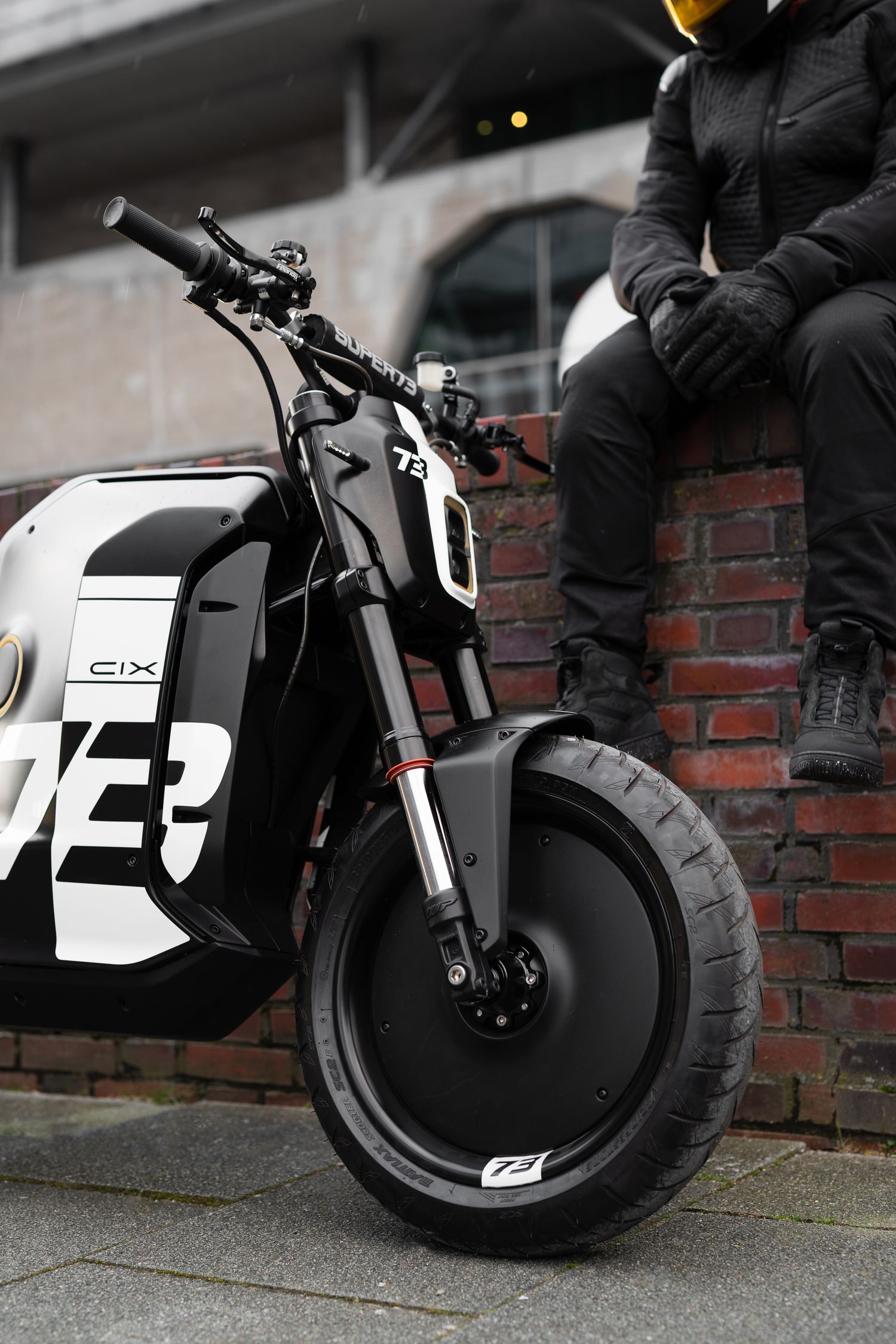 White and black electric motorcycle with distinctive wheel design against brick wall.