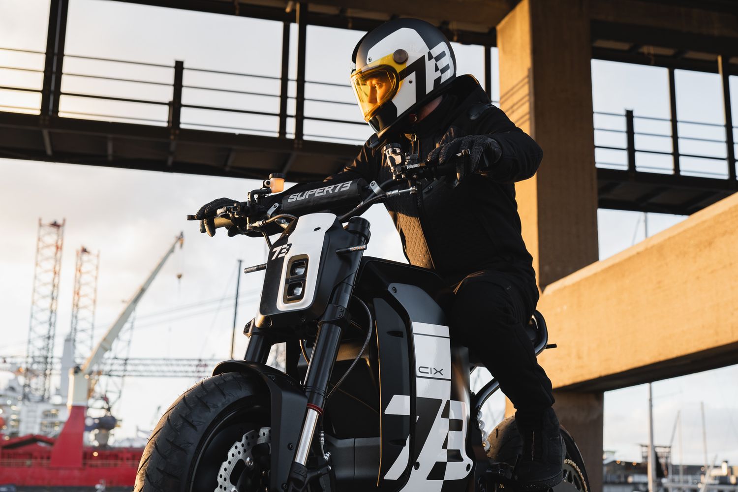 Silhouetted motorcycle against industrial skyline with cranes visible in background.