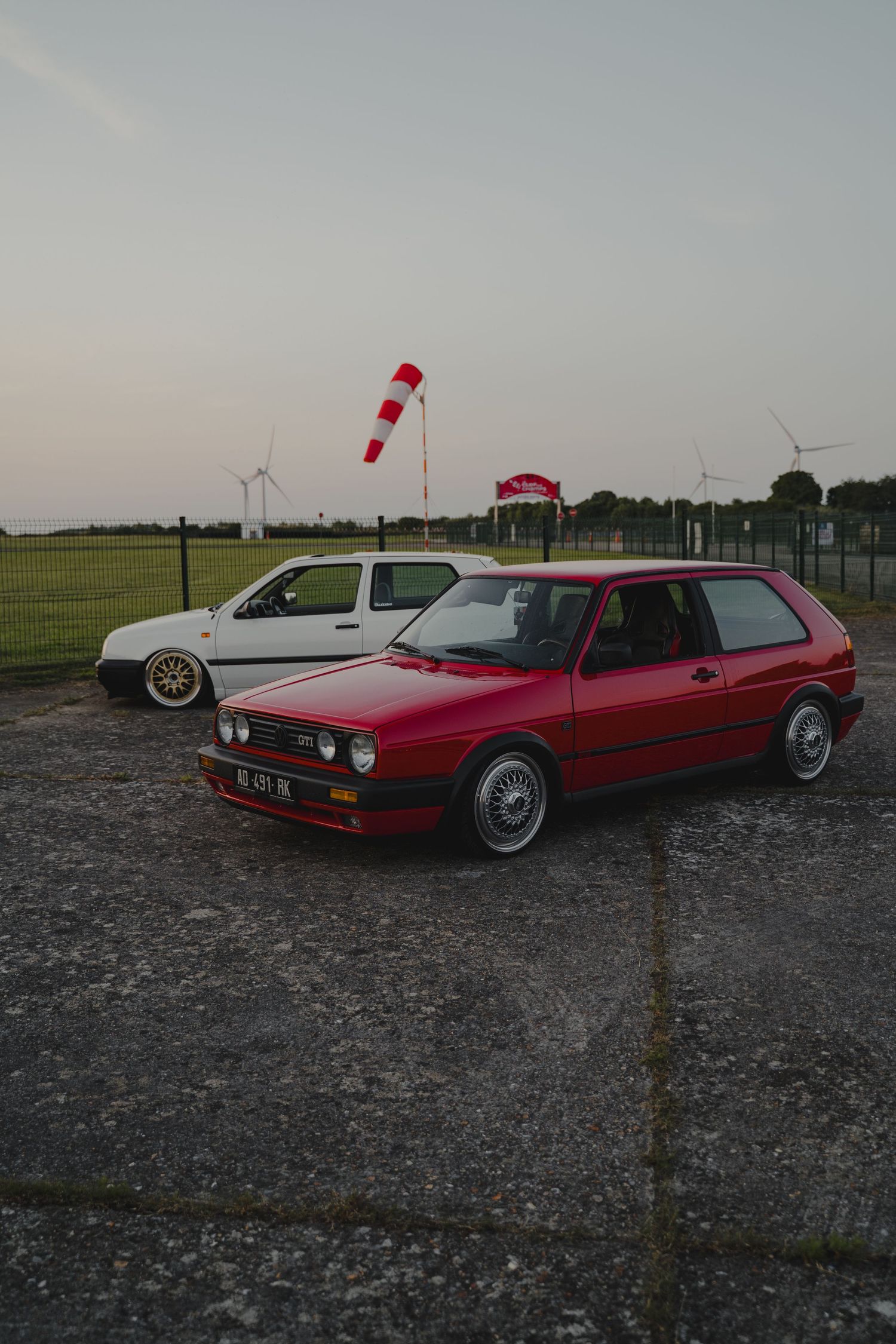 Row of vintage red cars parked on gravel with wind turbines in the background.