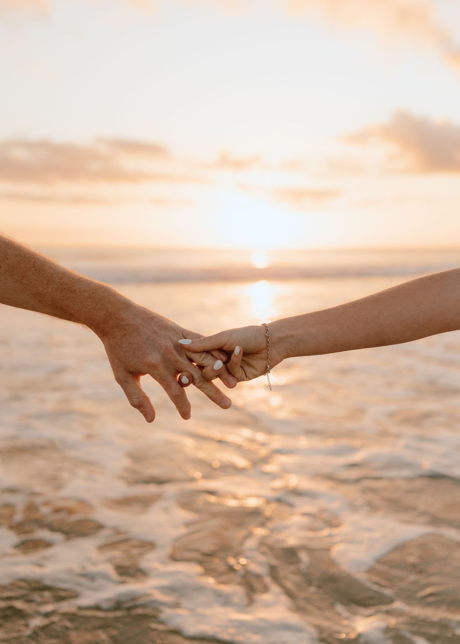 Two hands reach towards each other against a golden sunset over ocean waves creating a romantic silhouette.