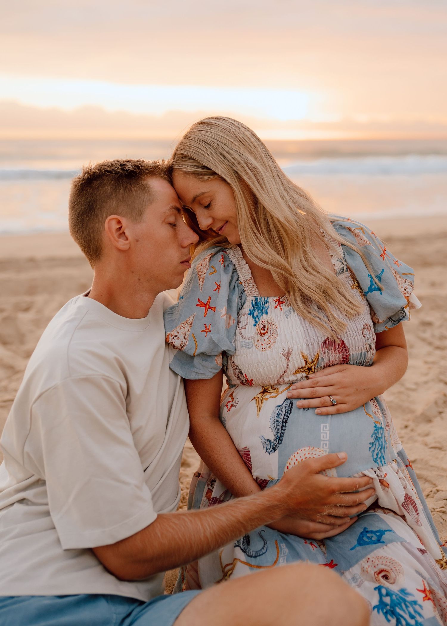 Couple shares tender moments on a beach at sunset in a romantic maternity photo series.