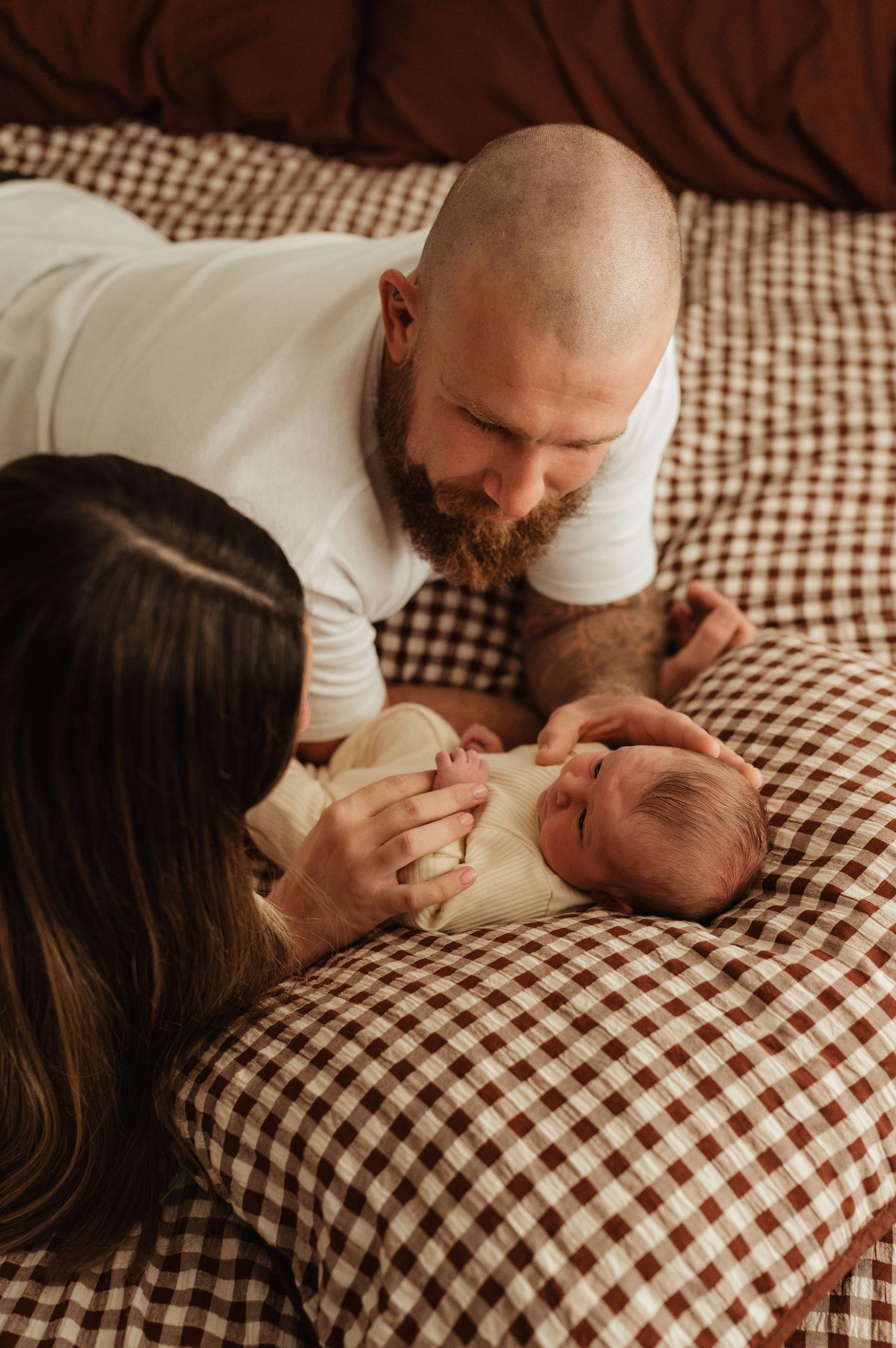 Parents lovingly gaze at their newborn baby on a brown gingham bedspread in a warm, intimate family portrait.
