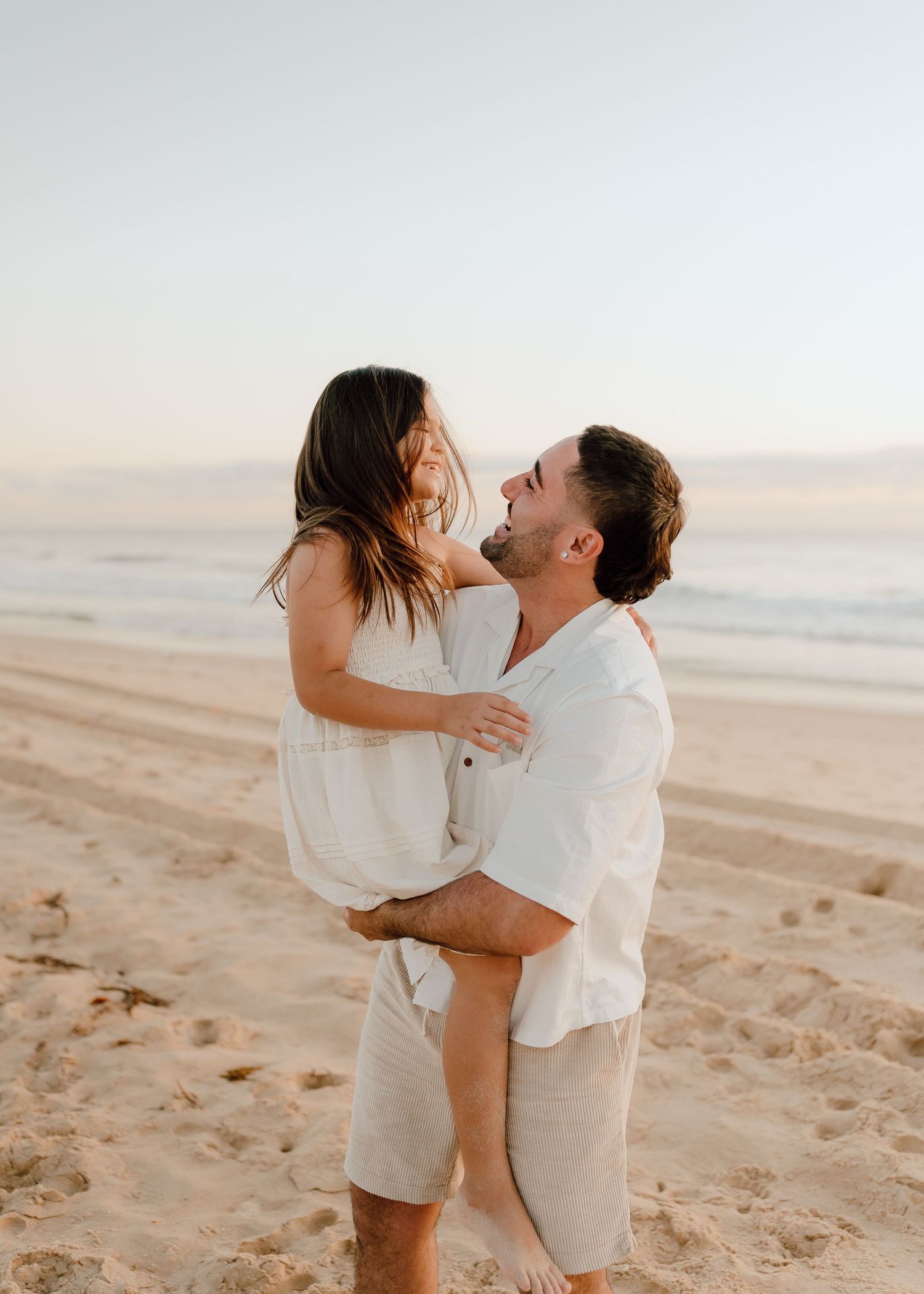 A couple embraces and laughs together on a sandy beach at sunset wearing white outfits.