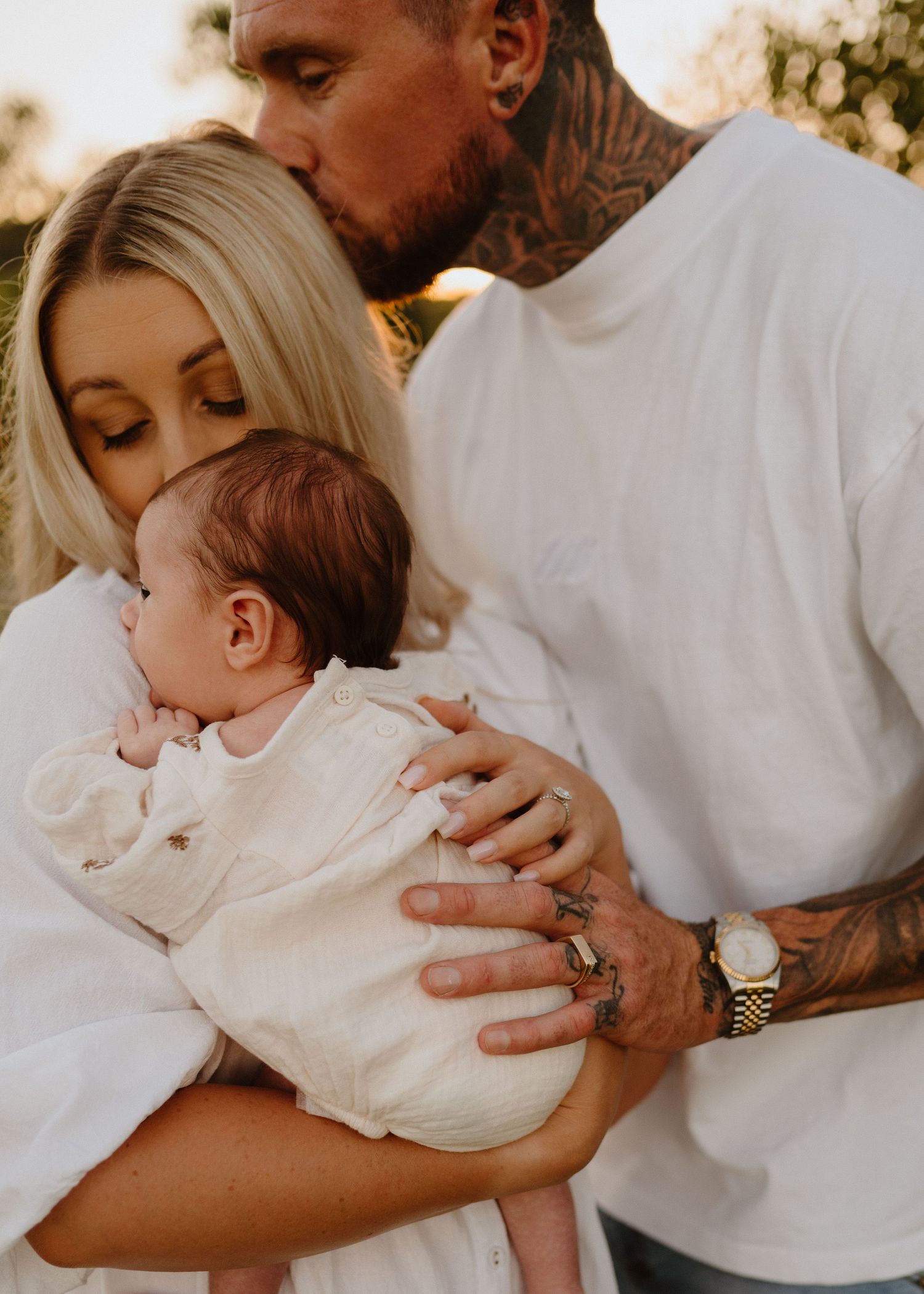 Tattooed father kissing blonde mother's head as they cuddle newborn baby in white outfits.
