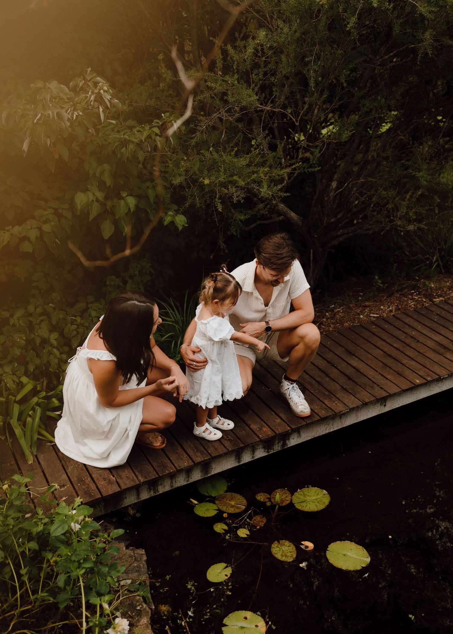Family of three sitting on a wooden bridge over a pond surrounded by lush greenery.