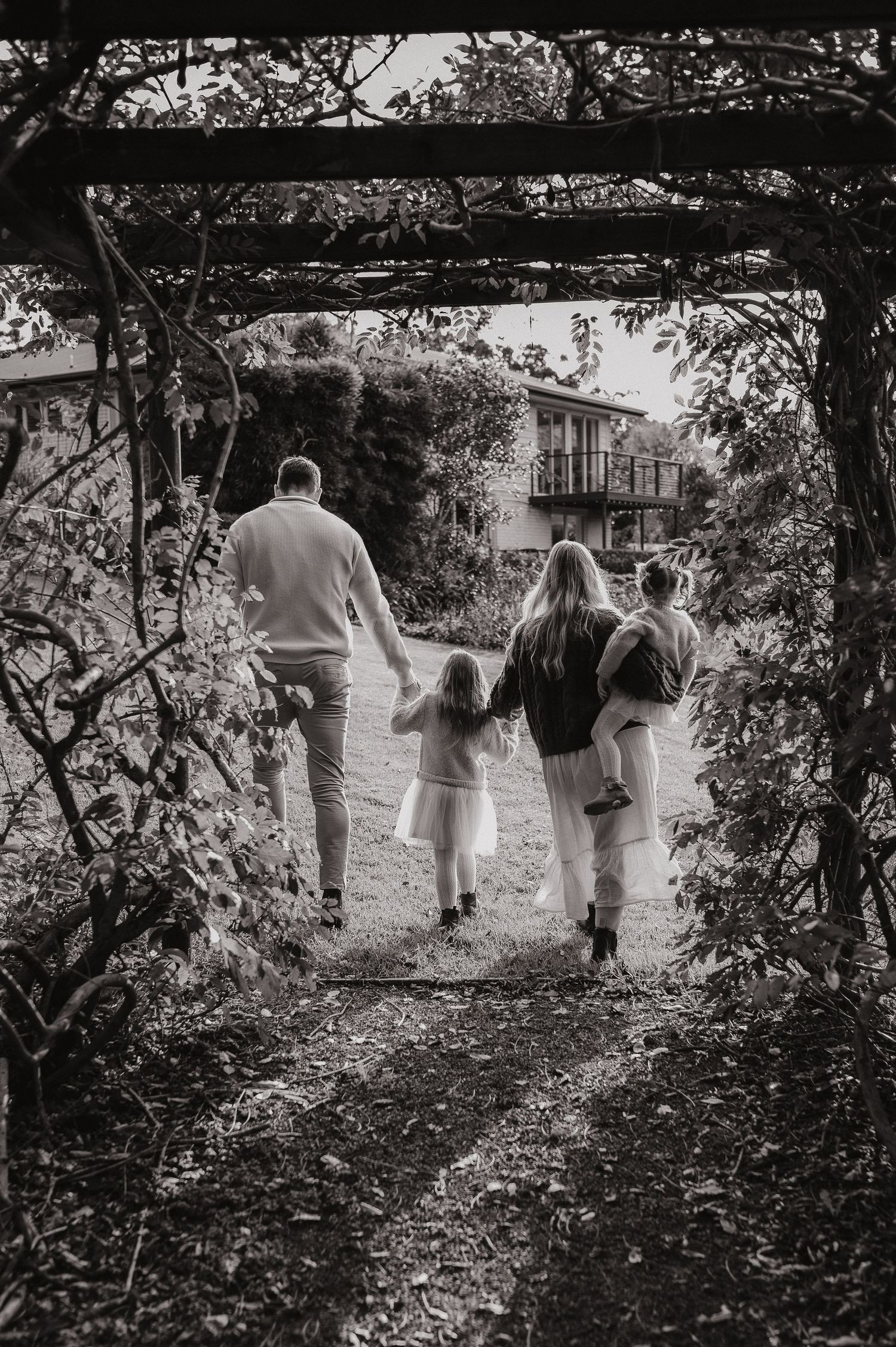 Black and white photo of a family walking hand-in-hand through a garden archway toward a house.