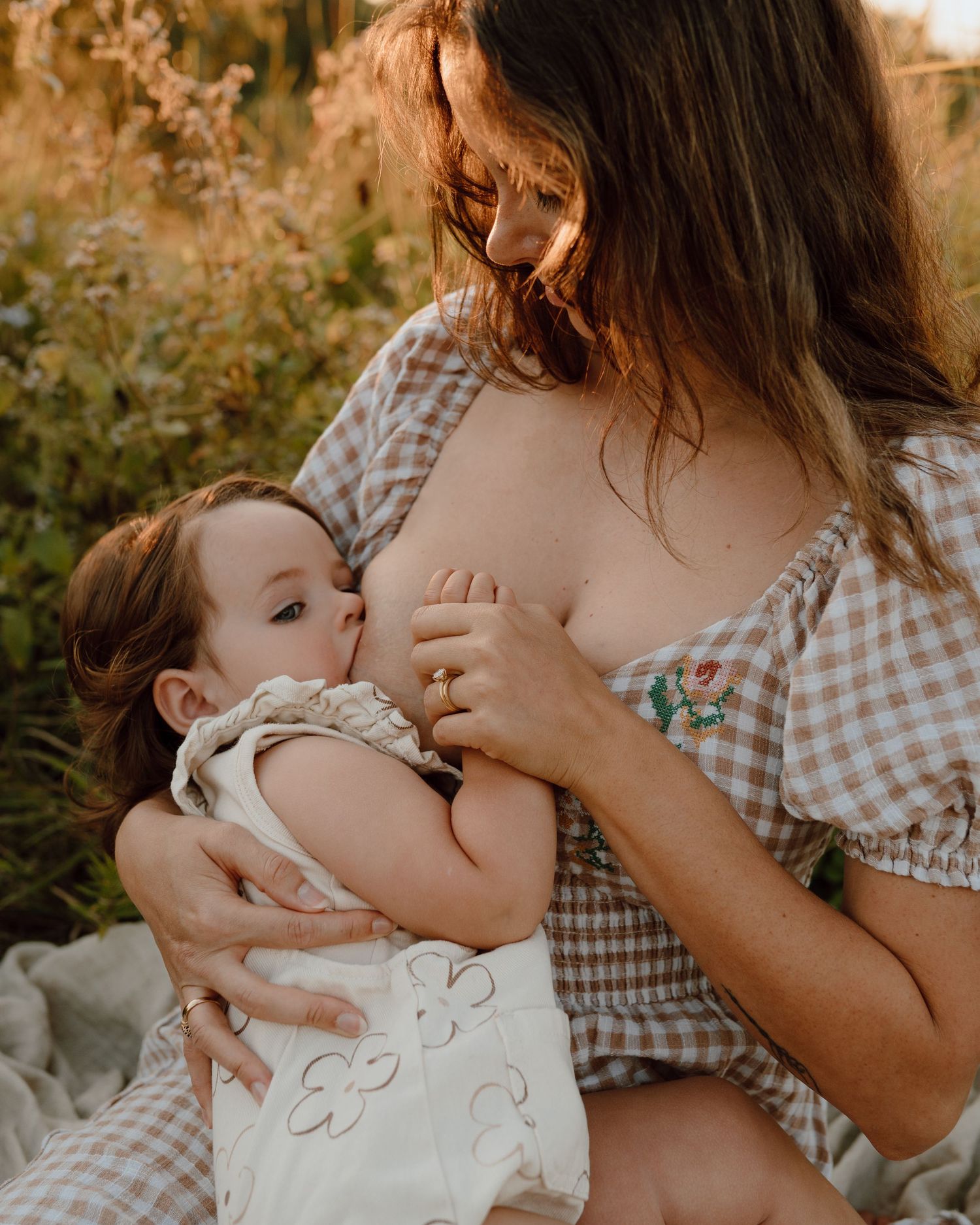 Mother nursing baby outdoors on blanket in gingham dress.