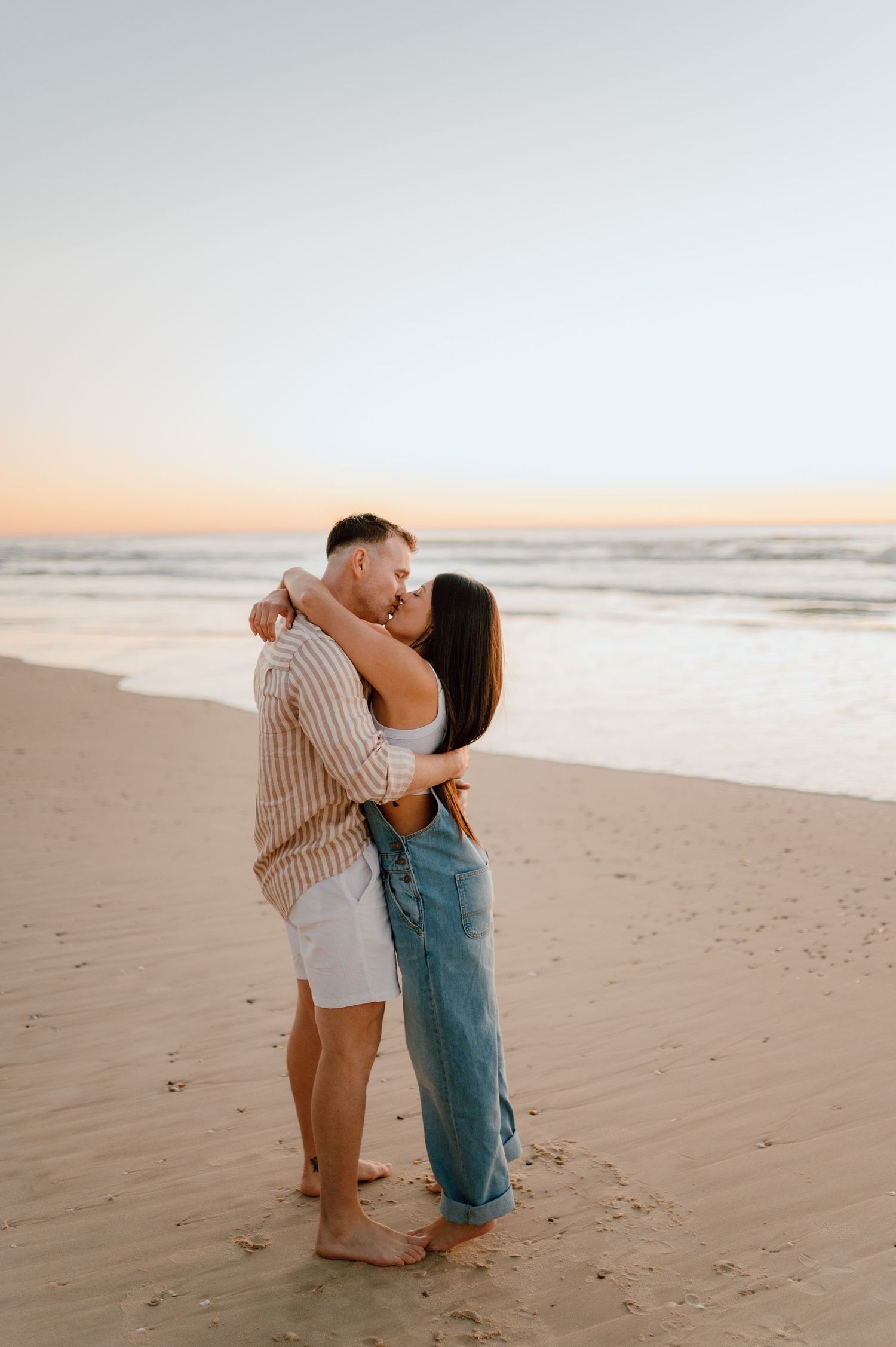 A romantic couple embraces on a sandy beach at sunset with ocean waves in the background.