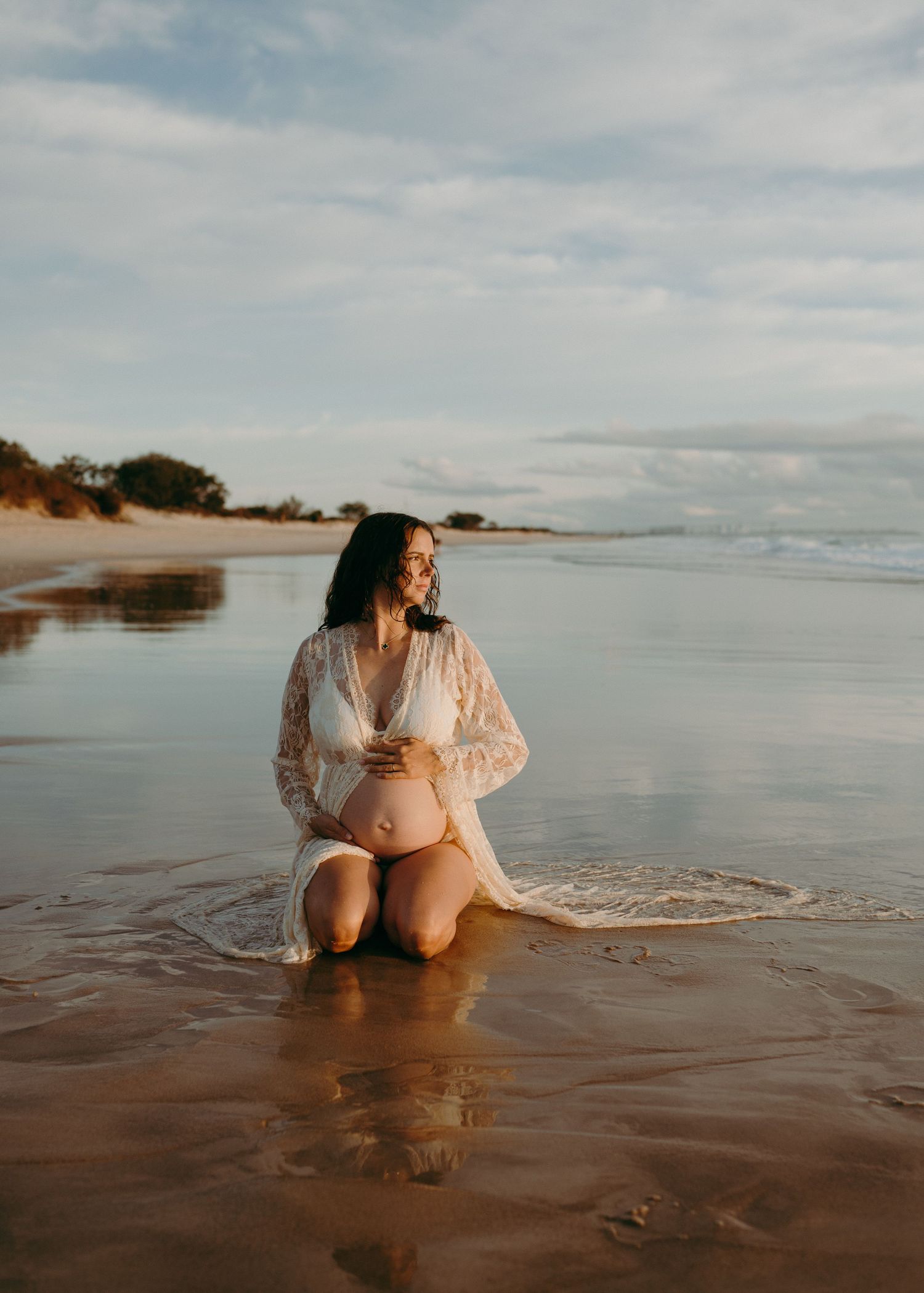 A pregnant woman in white lace dress sits at the edge of ocean waves during sunset on a sandy beach.