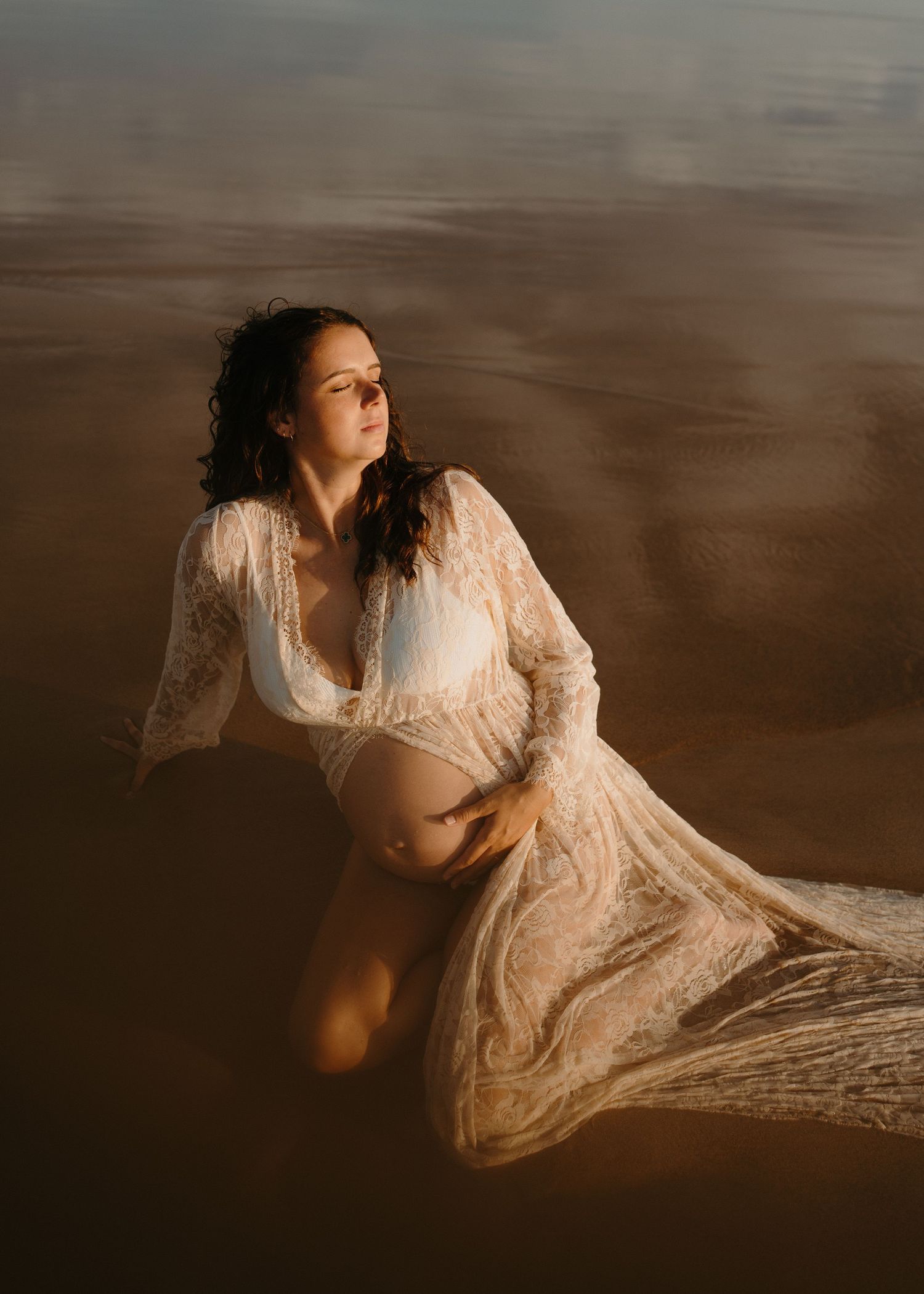 A person in a delicate lace dress poses gracefully on wet sand at the beach during golden hour.
