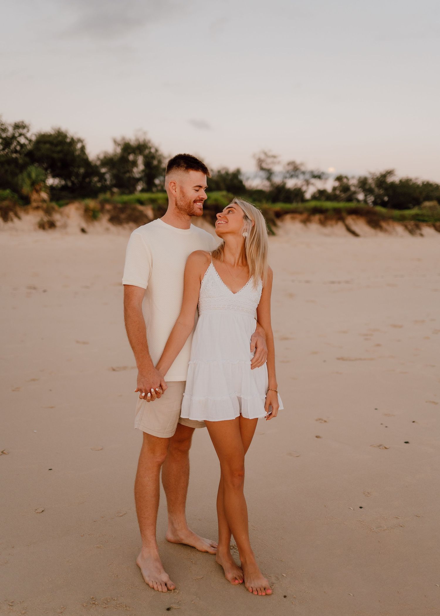A couple walking hand in hand on a sandy beach at sunset wearing white casual outfits.