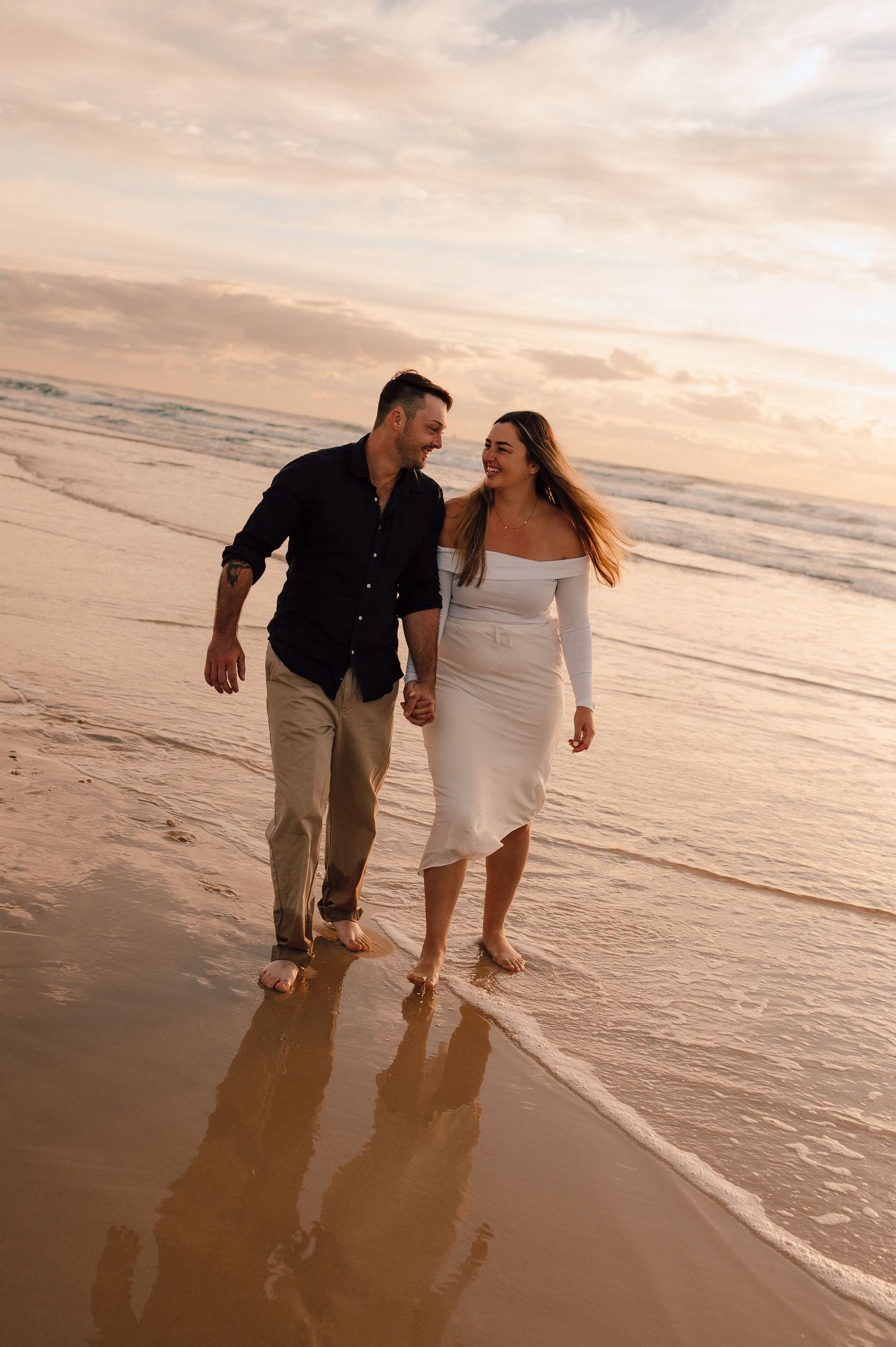 Couple walking hand-in-hand barefoot on a sandy beach at sunset, smiling at each other in casual and white dress attire.