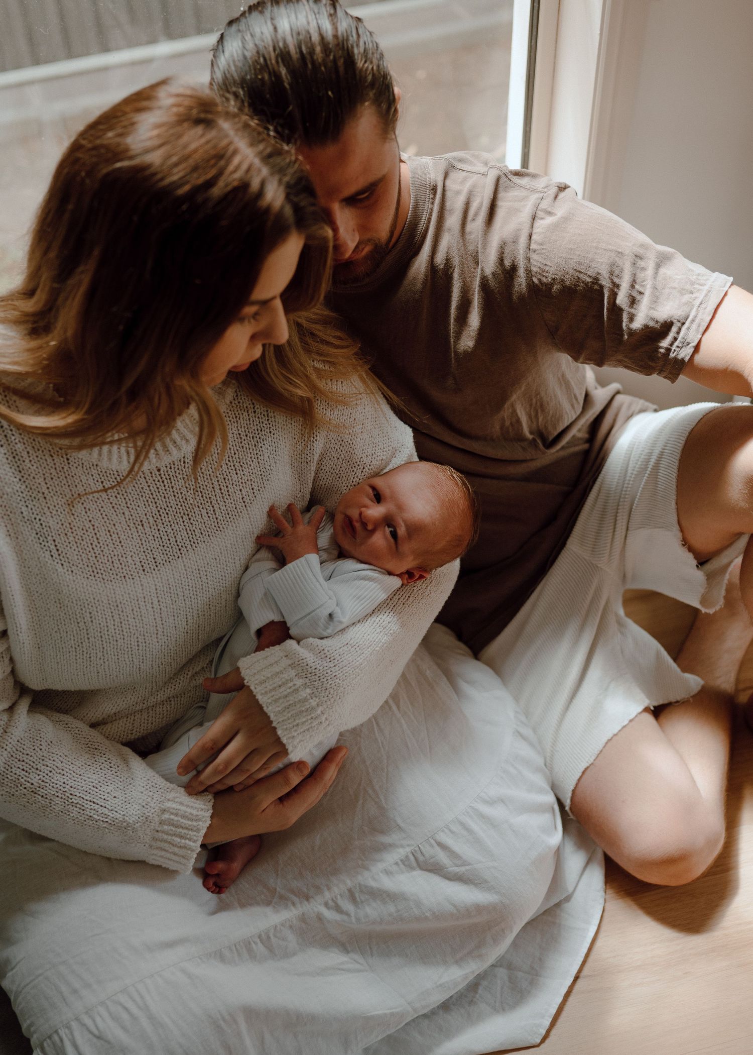 Family shares tender moment together while sitting on white bedding in natural light.