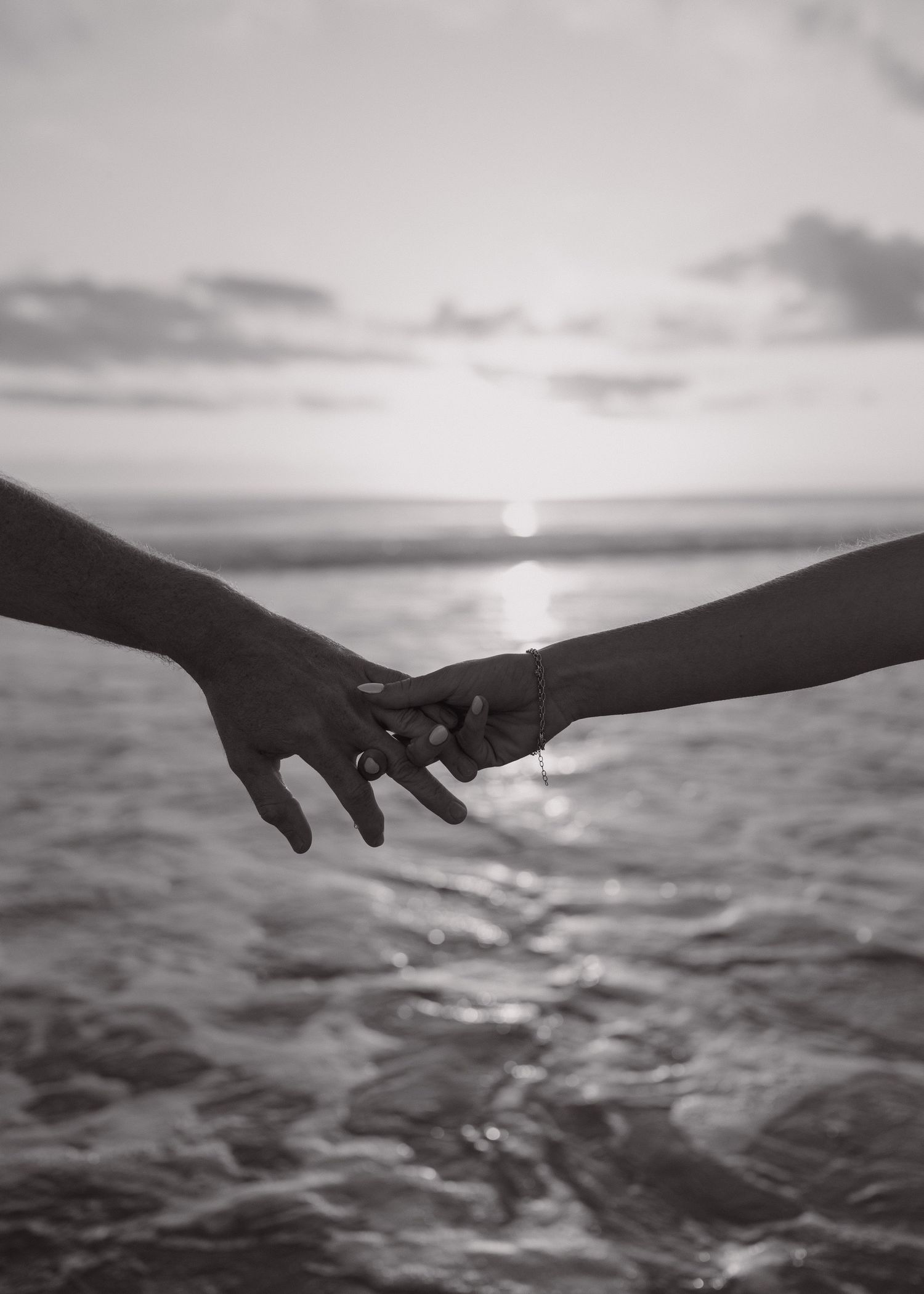 A black and white photograph shows a silhouetted hand reaching towards a sunset over a serene ocean horizon.