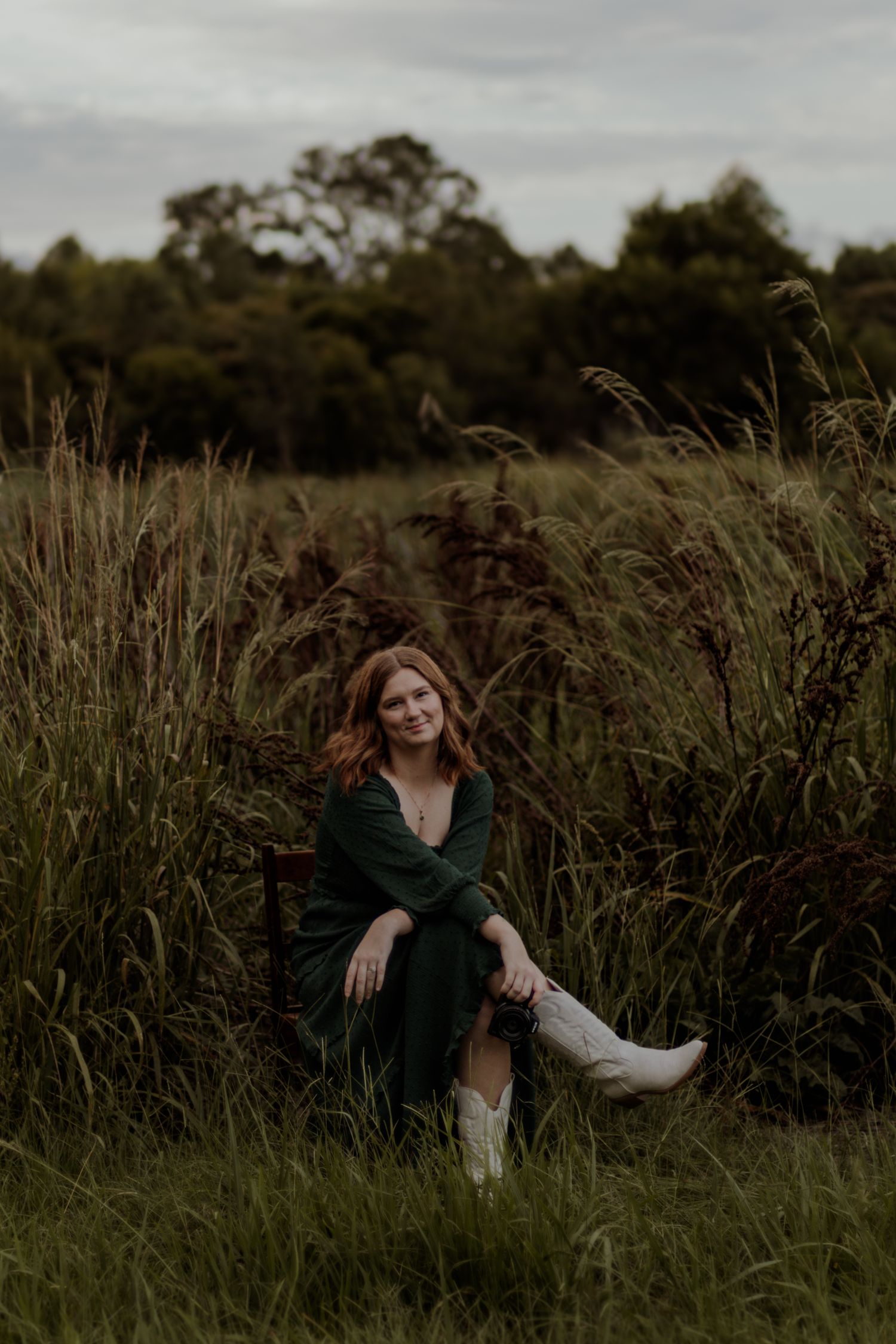 A woman in a dark dress poses gracefully among tall grass in a moody outdoor setting at dusk.
