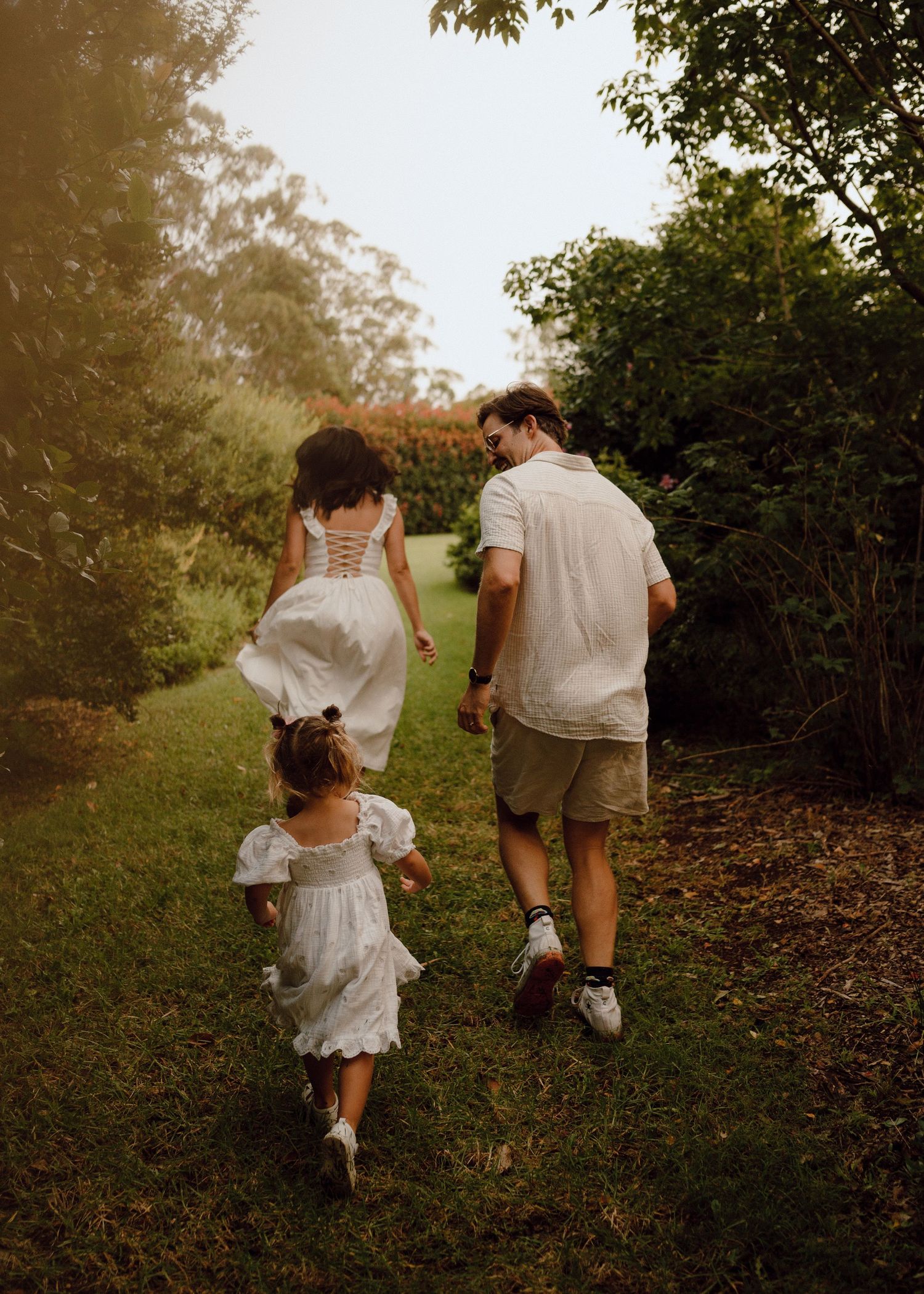 Family of three walking hand in hand through a lush garden path, dressed in coordinating white outfits.