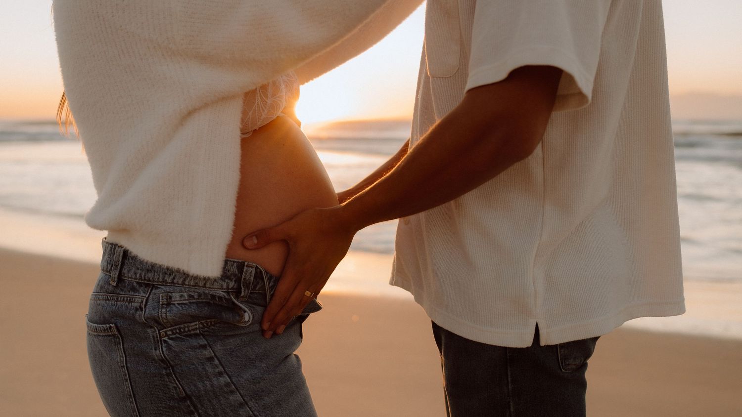 Couple holding hands on a beach at sunset, wearing white shirts and jeans, with golden light reflecting on the ocean.
