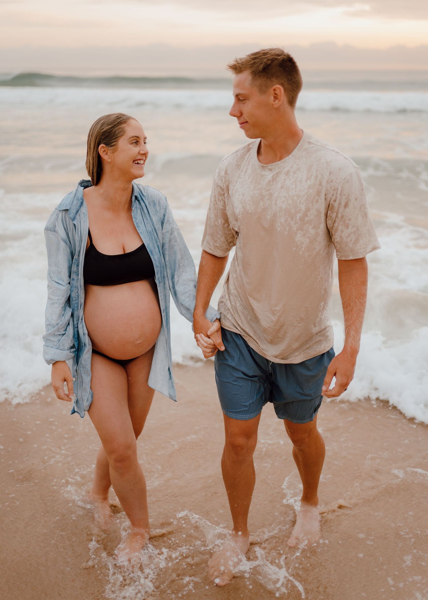 A couple walks hand in hand along the beach at sunset, splashing through gentle waves in casual summer attire.