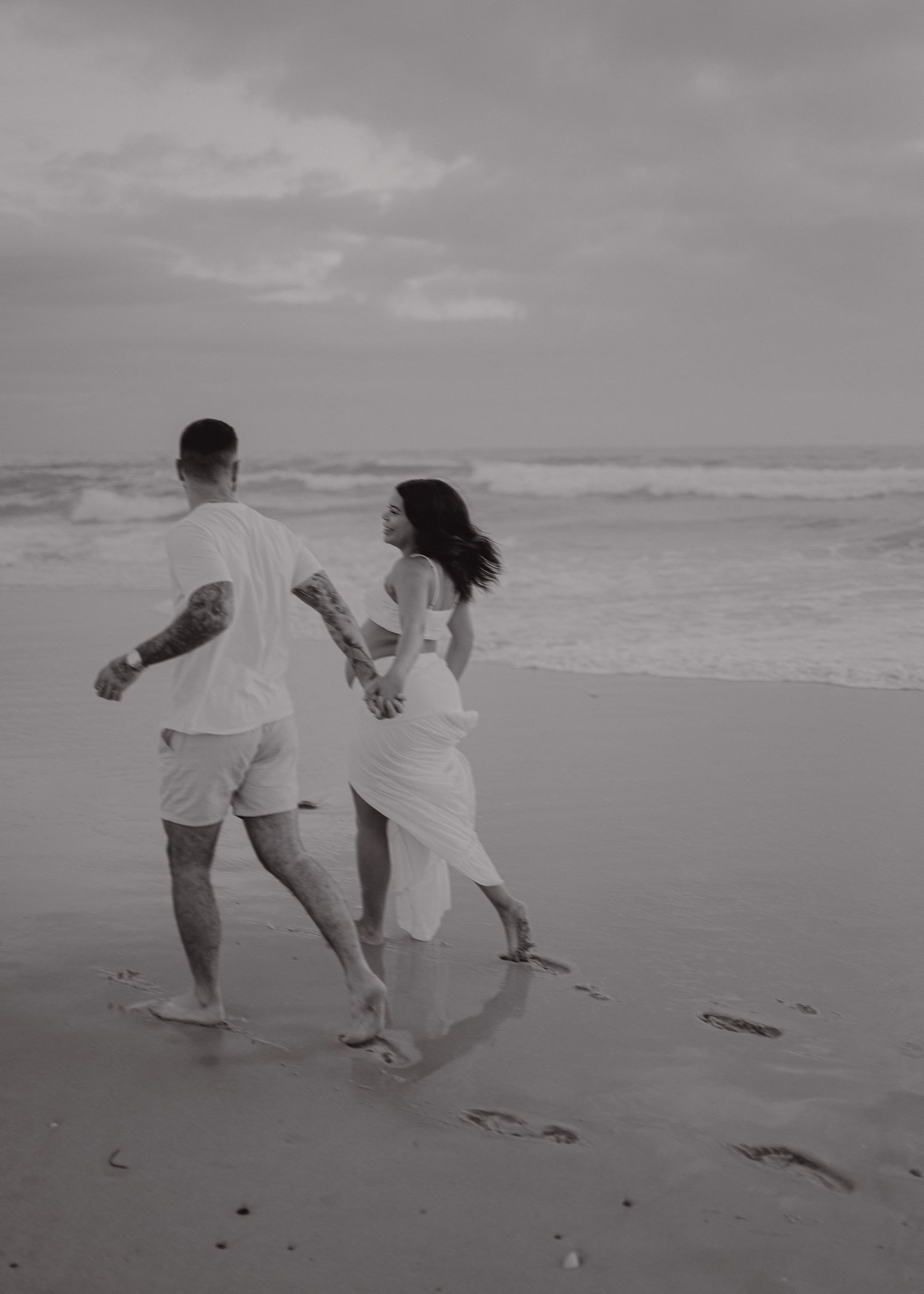 A couple in white outfits runs joyfully along the beach holding hands, leaving footprints in the sand.