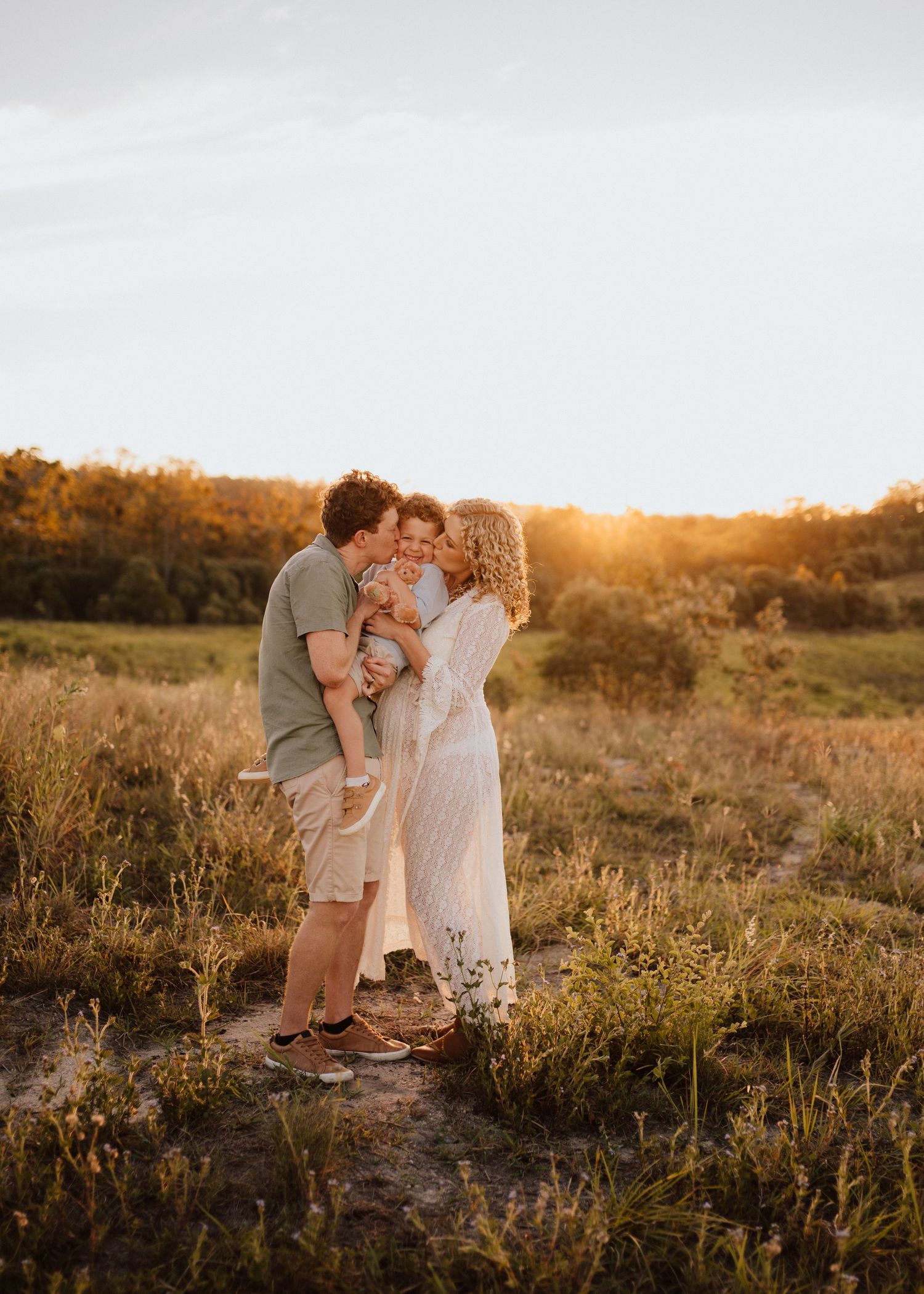 A couple shares a tender moment in a sunlit field during golden hour as their child embraces them.