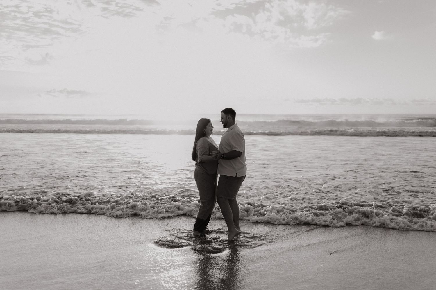 Black and white photo of a couple embracing on a beach with ocean waves behind them.