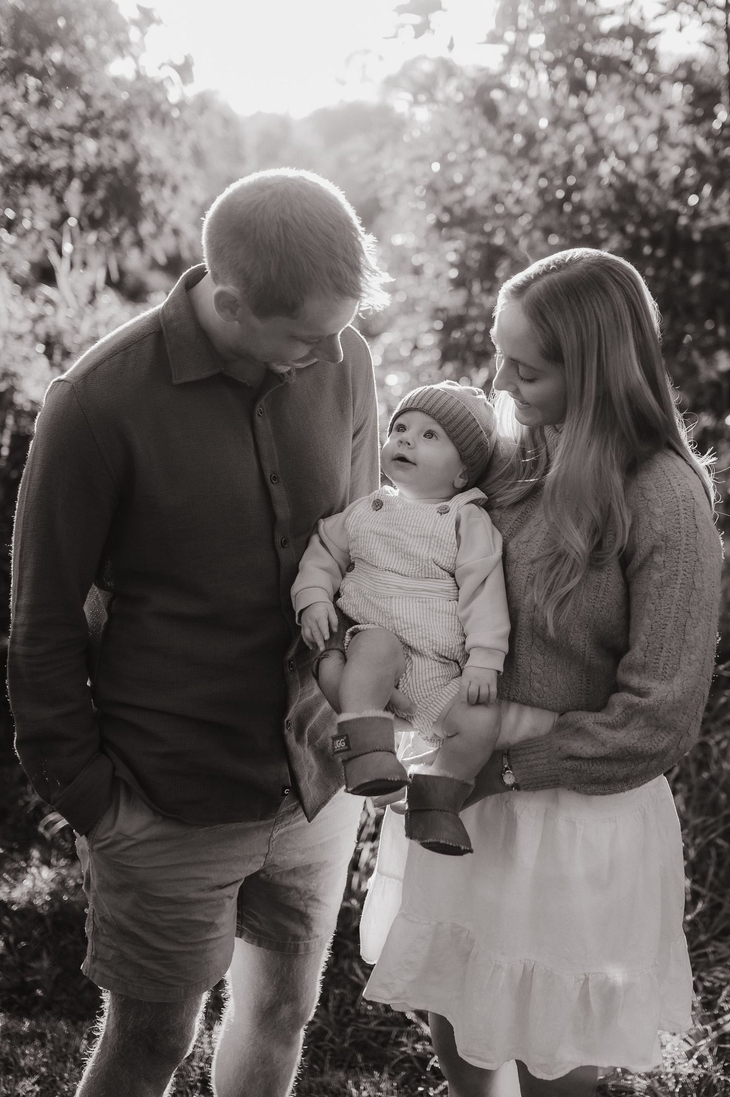 Black and white photo of a young couple smiling at their baby outdoors among trees.