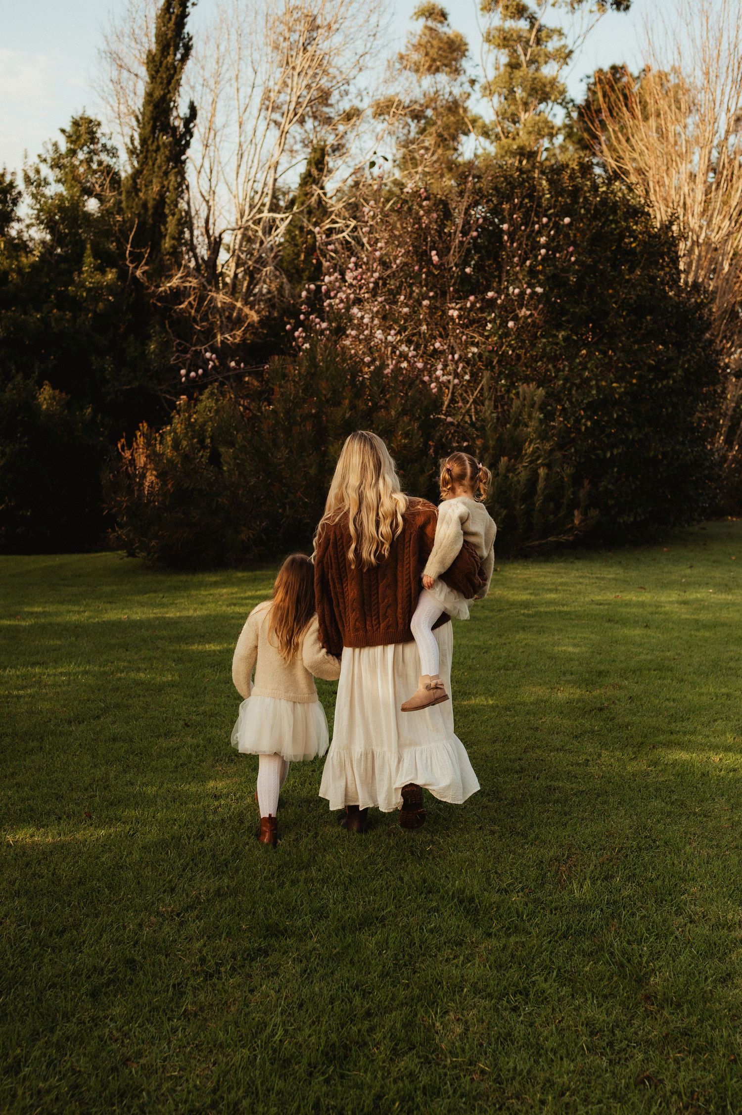 A mother walks with two young daughters across a green lawn toward lush garden trees in warm golden light.