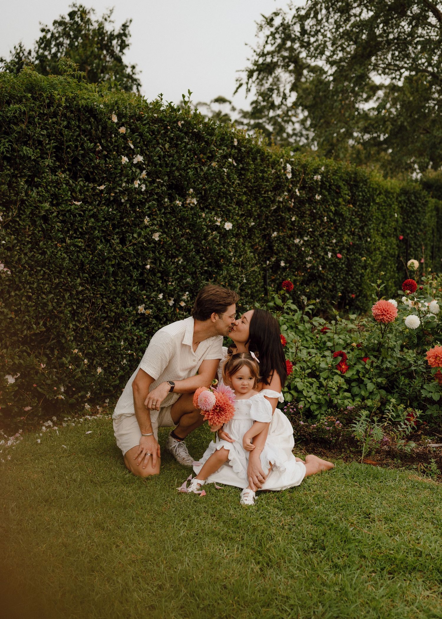 Parents kissing their toddler daughter in a rose garden, all dressed in white on a green lawn.