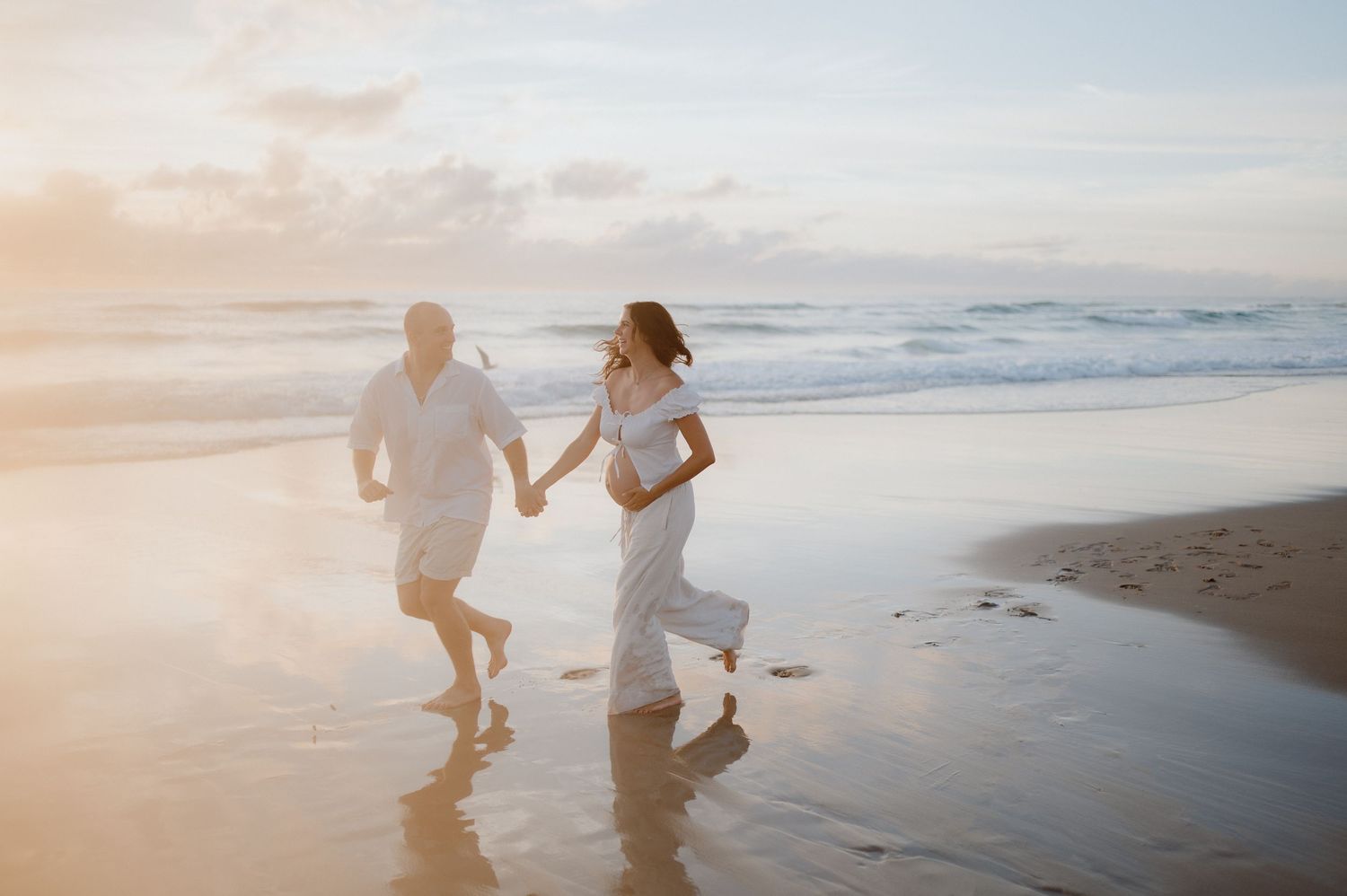 A couple walks hand in hand along a misty beach at sunset with soft waves rolling onto the shore.