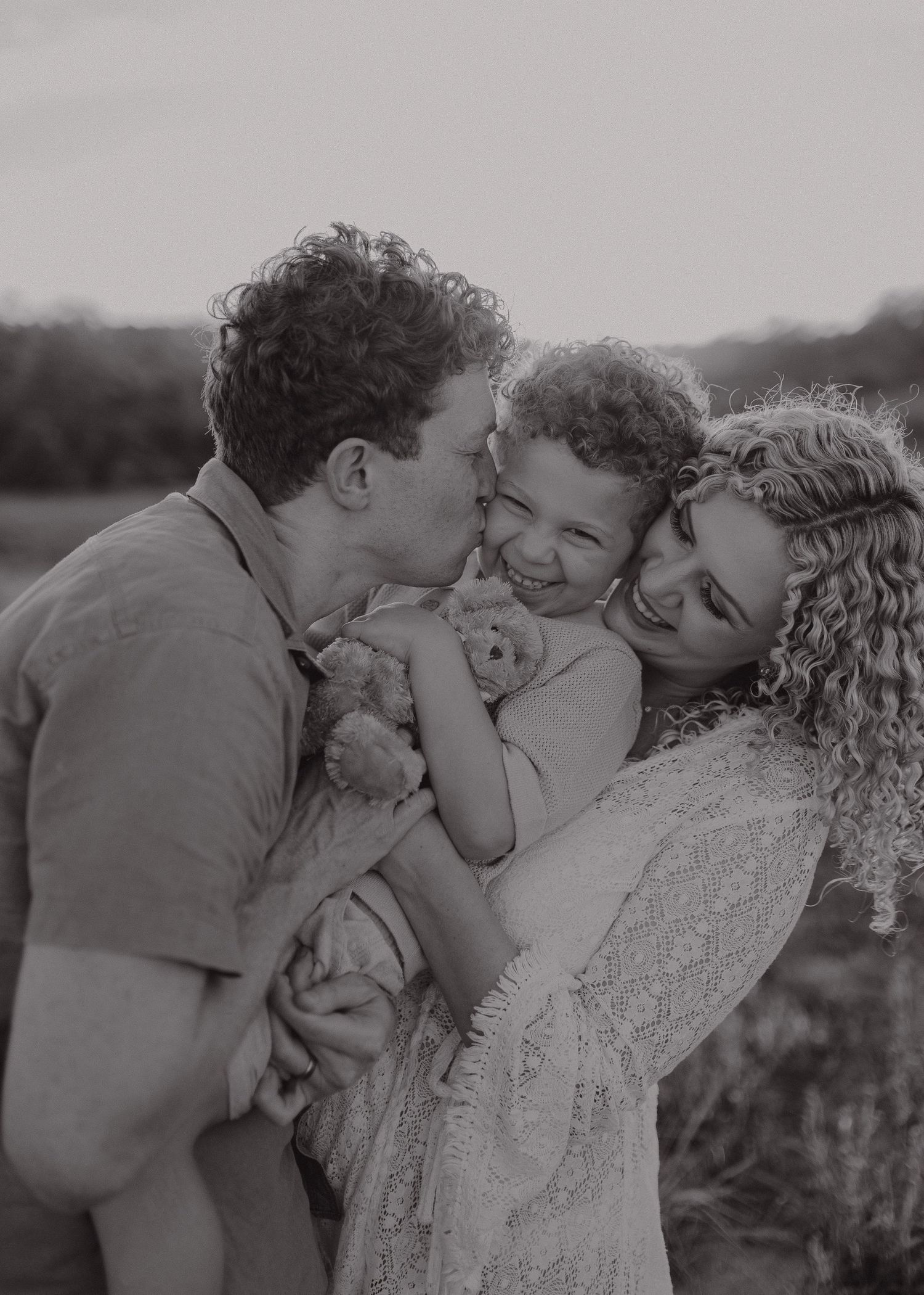 A tender family moment captured in black and white as they share kisses and cuddles outdoors in natural light.