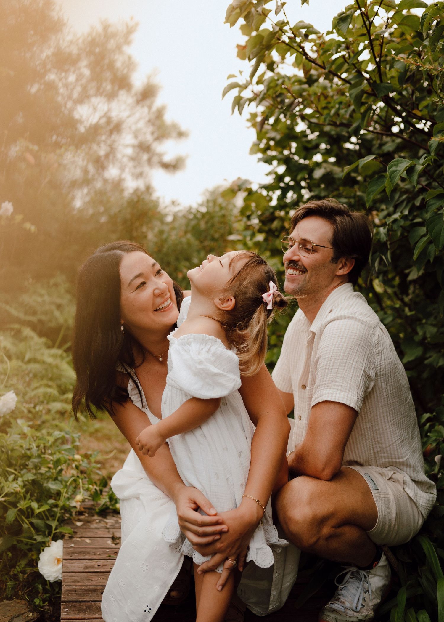 Family of three laughing together outdoors in a lush garden, parents hugging their young daughter in white dress.