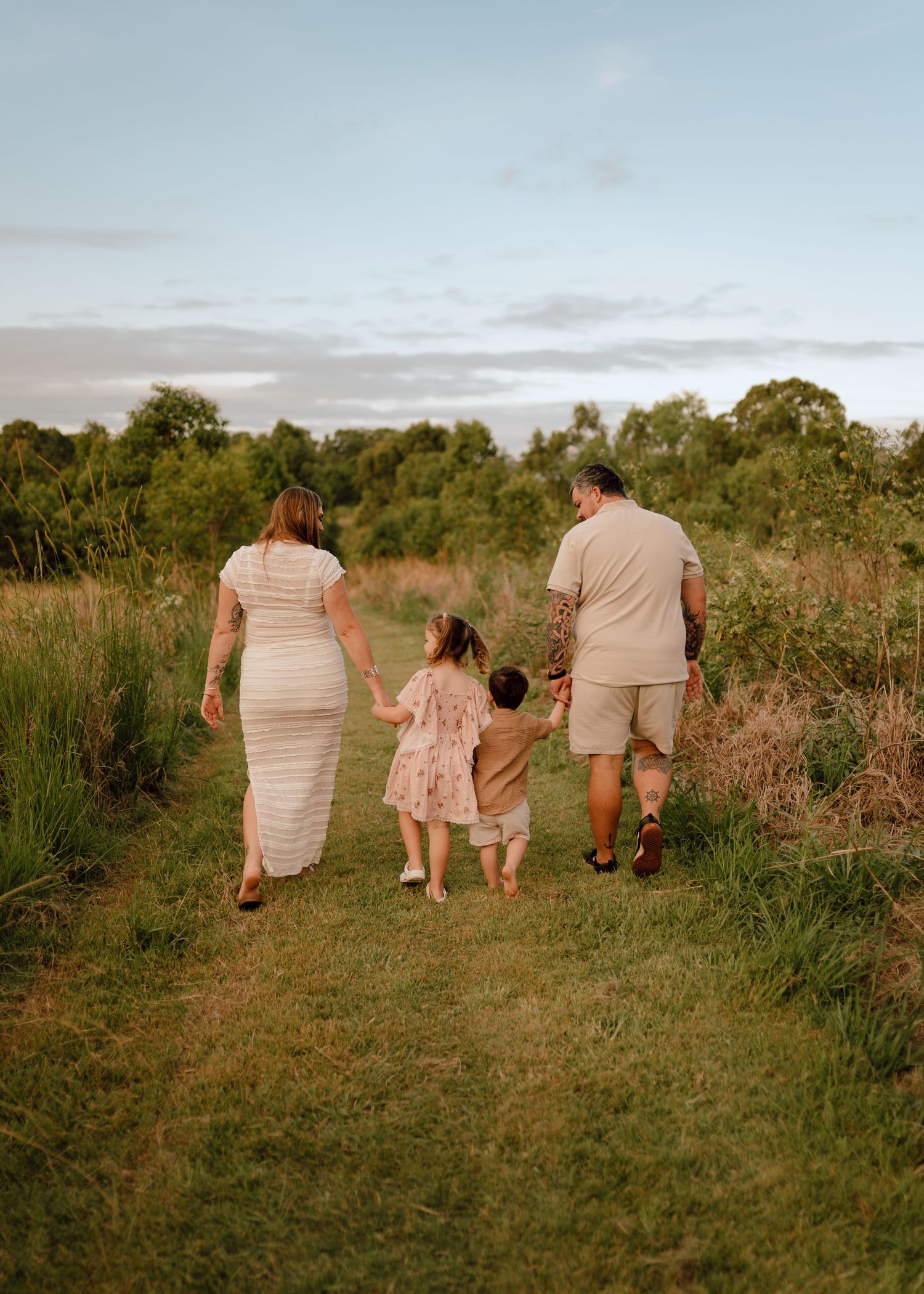 A family walks together on a grassy path through tall grass at sunset, viewed from behind.