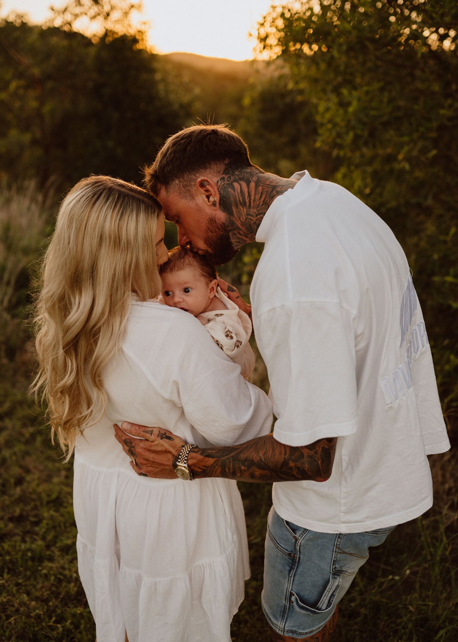 A tattooed couple in white outfits shares a tender moment kissing their newborn baby outdoors at golden hour.