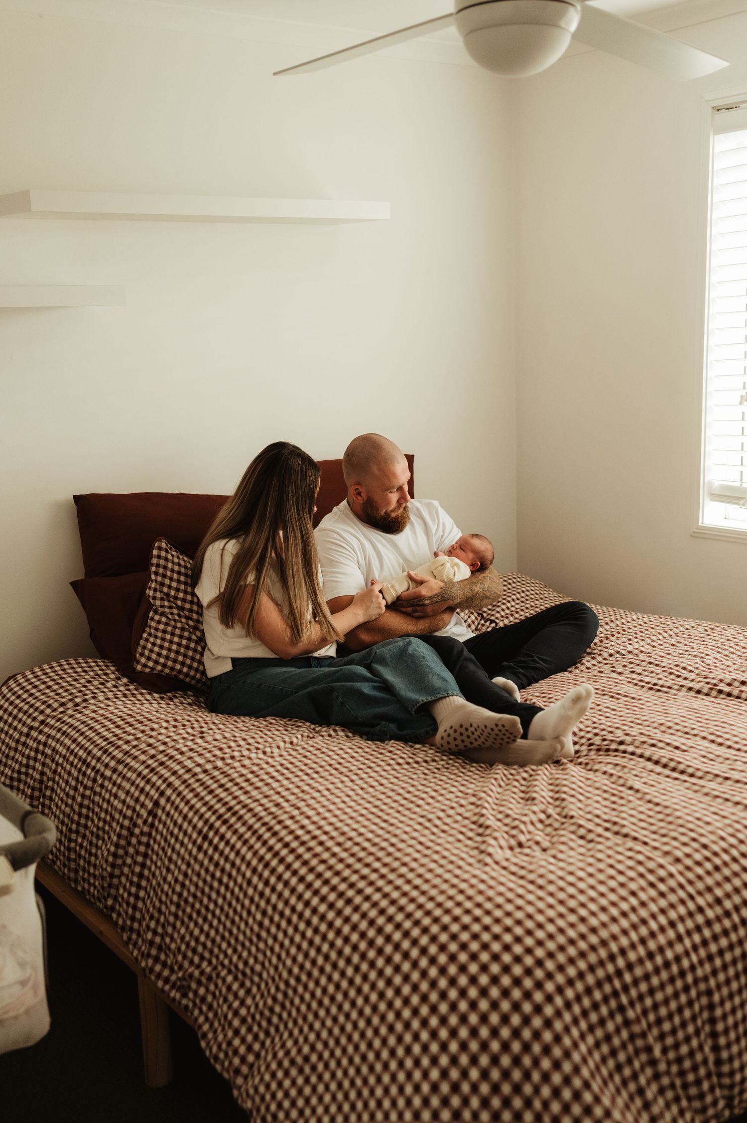 Couple sitting on a gingham bed holding their newborn baby in a cozy bedroom.