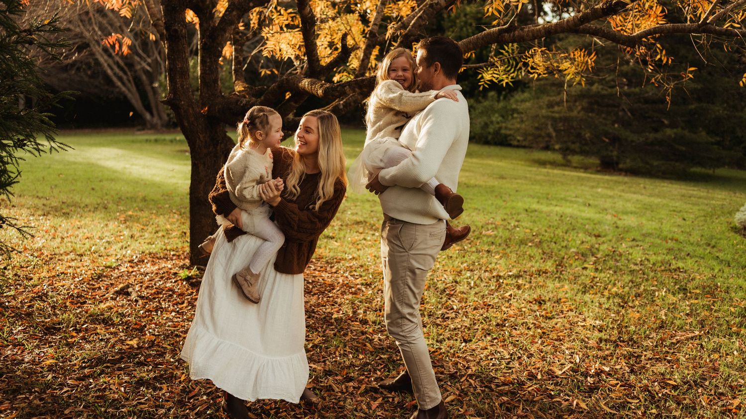 Family of four enjoying autumn park, parents holding toddlers among golden fallen leaves near a large tree.