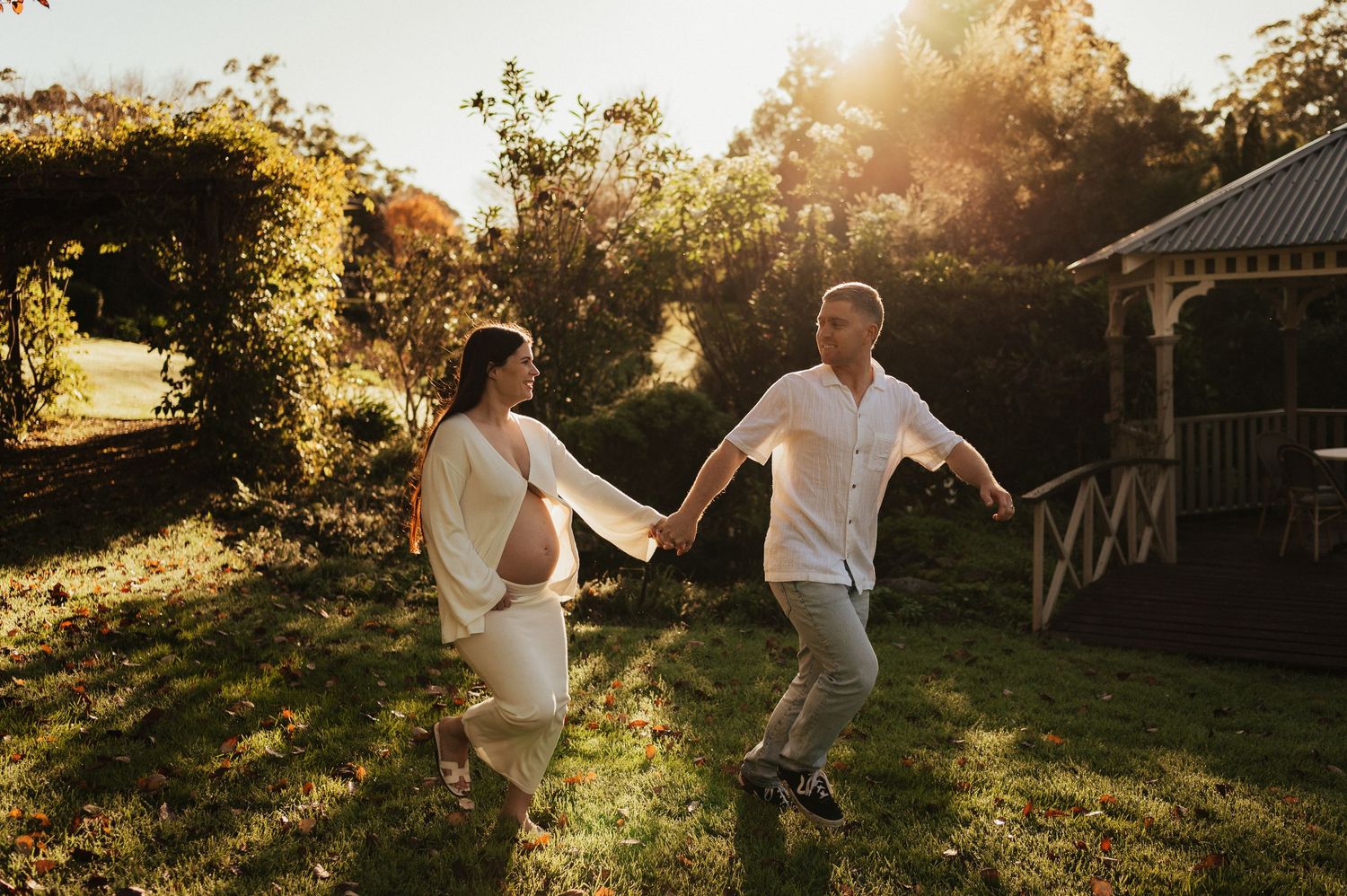 Pregnant woman and man holding hands dancing in sunlit garden near gazebo during golden hour maternity photoshoot.