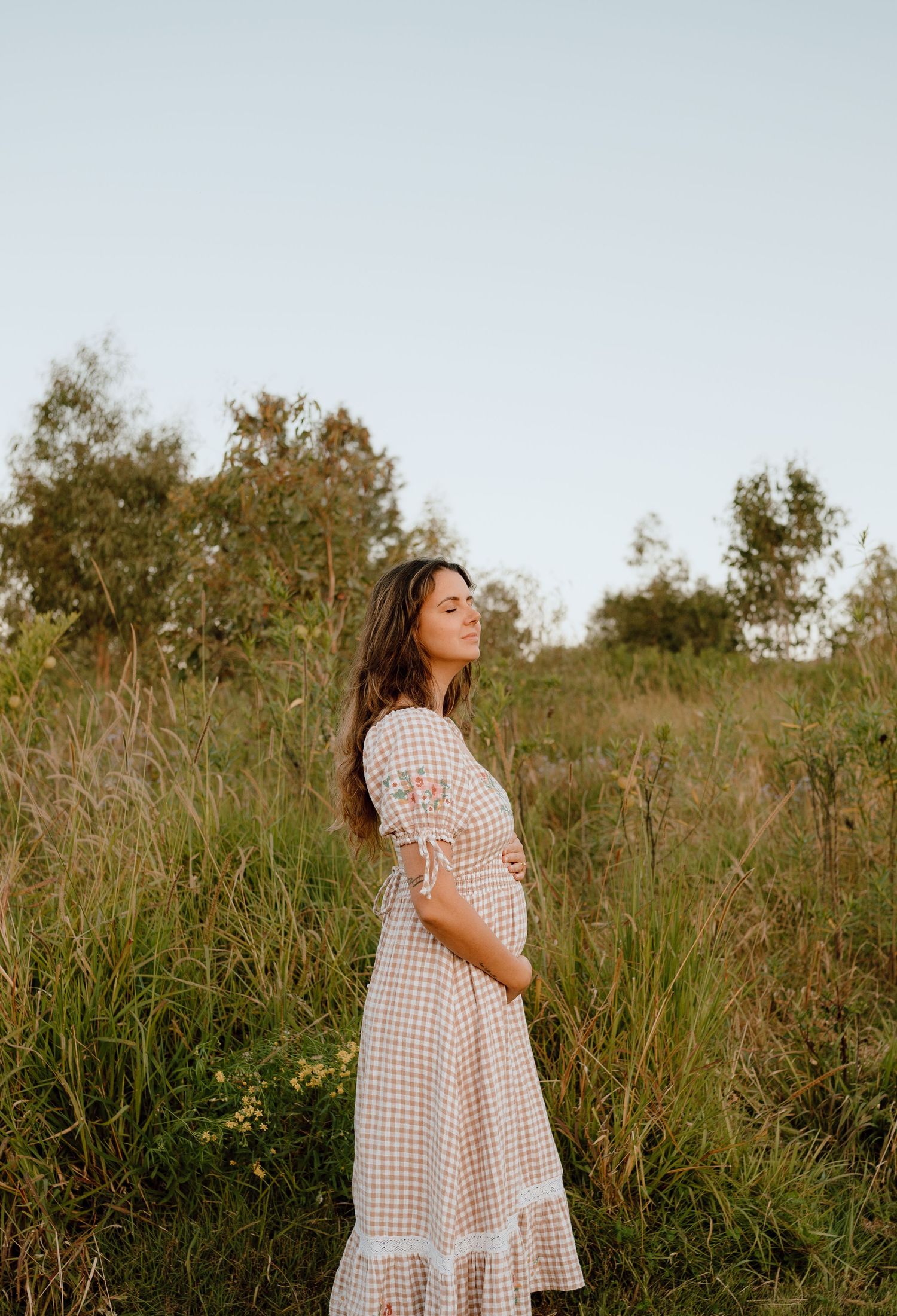 Woman in gingham dress stands in golden field during sunset.