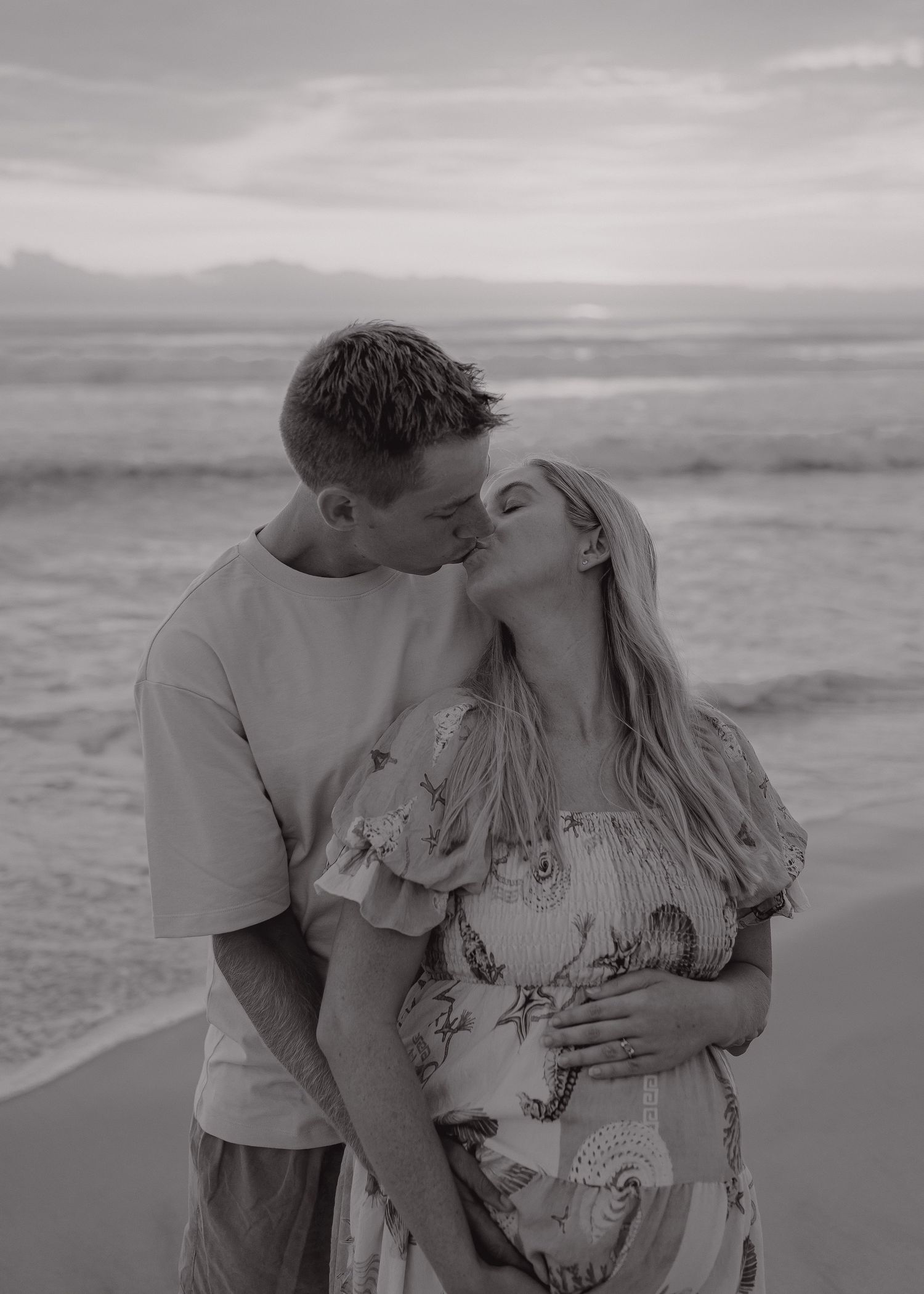 A couple shares a romantic moment at the beach in black and white, with the ocean waves visible in the background.