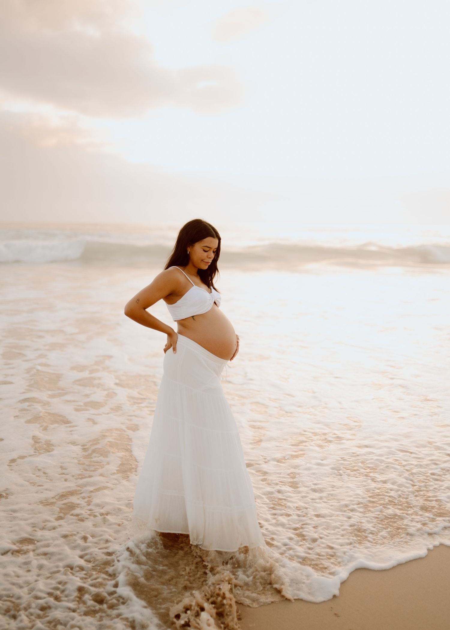 Pregnant woman in white dress walking along beach shoreline at sunset creating artistic silhouette photos.