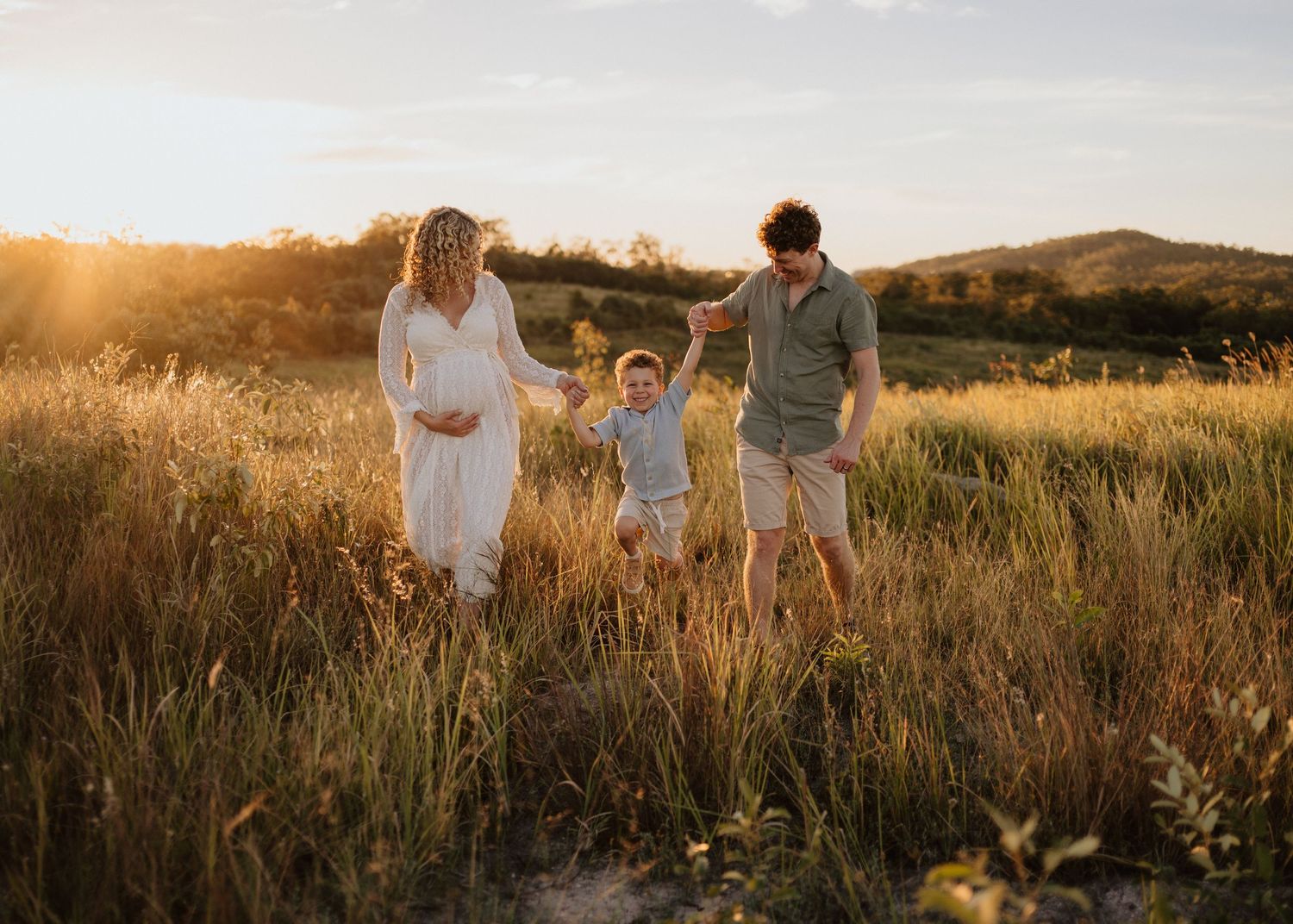 A family walks together through a sunlit meadow during golden hour as warm light illuminates the tall grass.