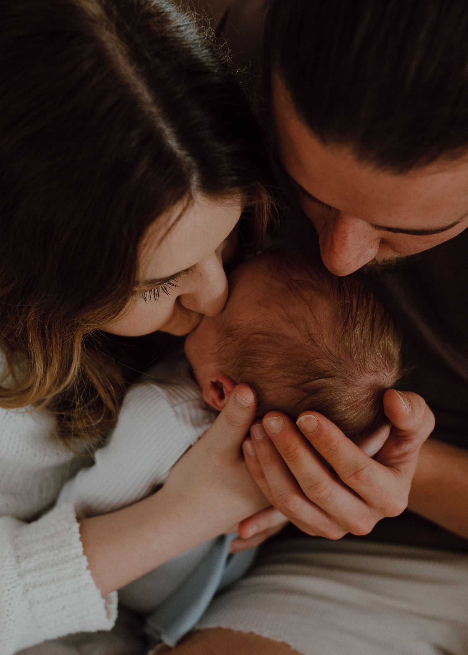 A tender moment as parents gently cradle and kiss their newborn baby in soft natural lighting.