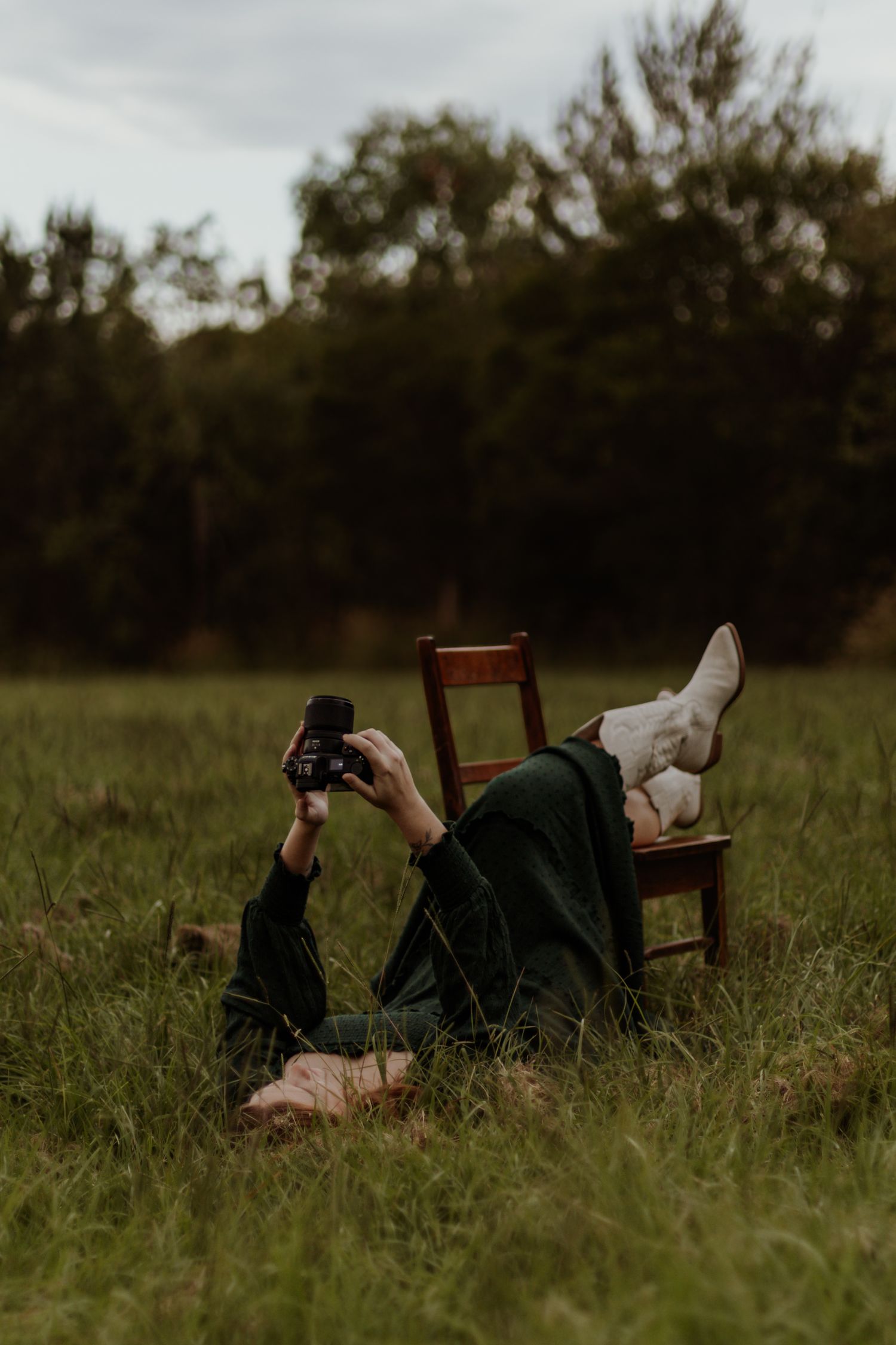 A photographer lies in tall grass next to a wooden chair while taking photos in a moody outdoor setting.