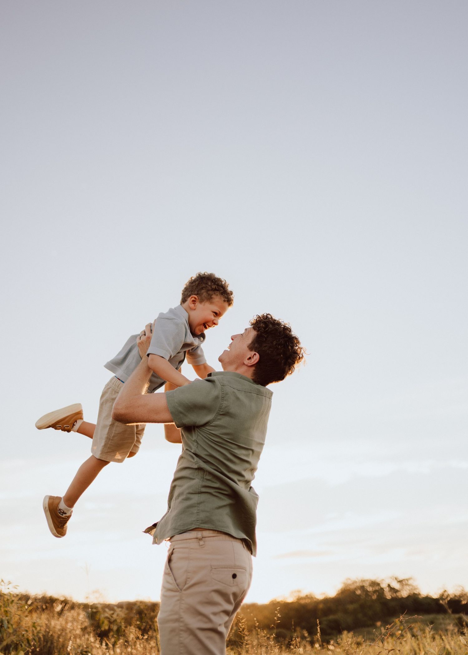 A parent lifts and plays with a small child outdoors during golden hour in a grassy field.