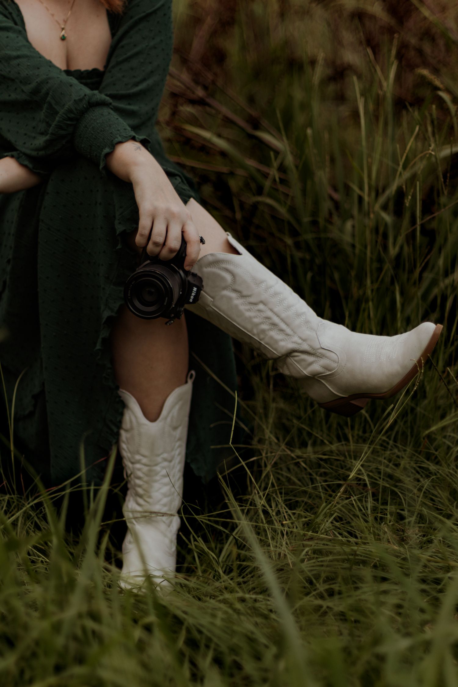 White cowboy boots with embroidered details stand out against dark grass in a rustic outdoor setting.