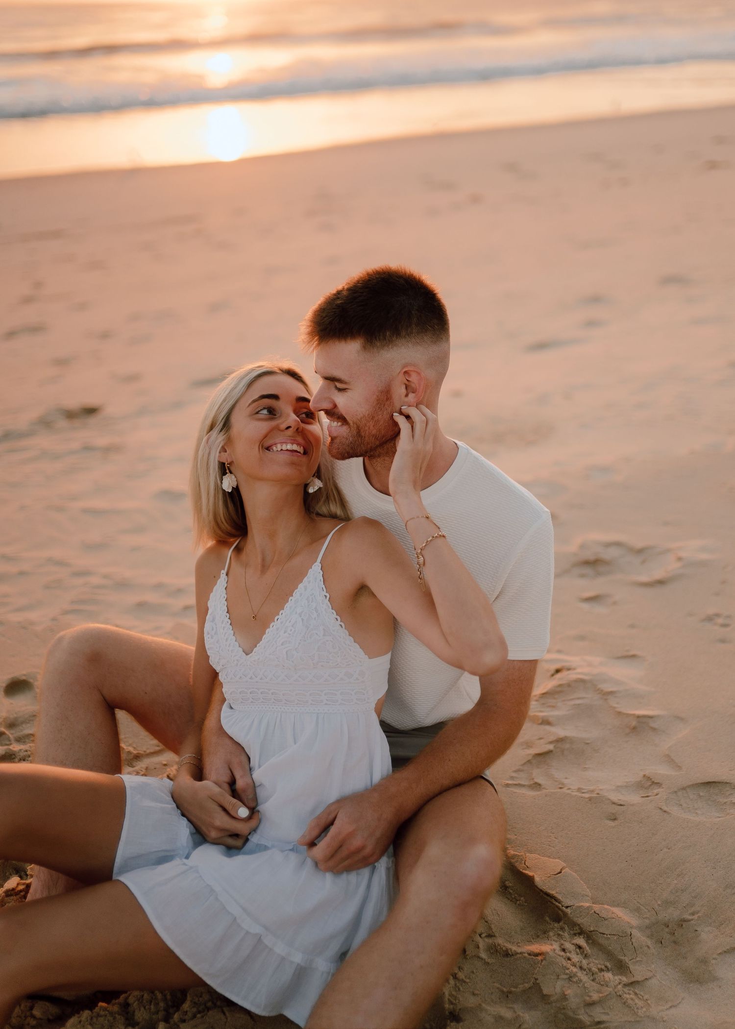 Couple in white outfits sharing intimate moments on a sandy beach during golden hour sunset.