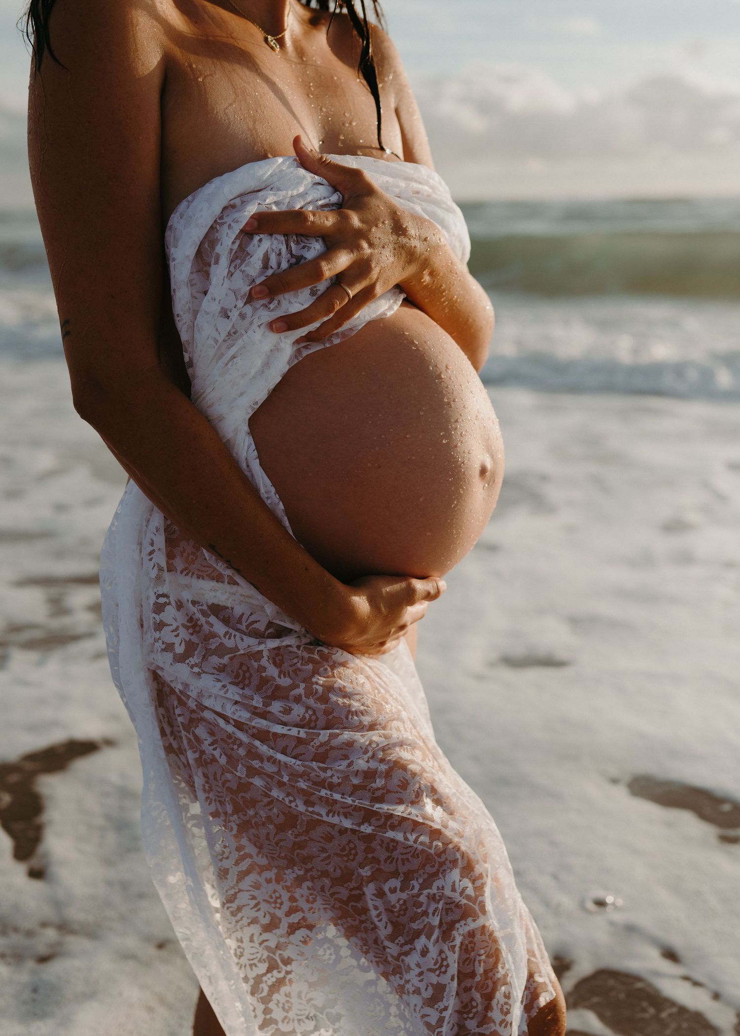 A silhouetted pregnant belly is photographed against a beach sunset with ocean waves and foam creating an artistic effect.