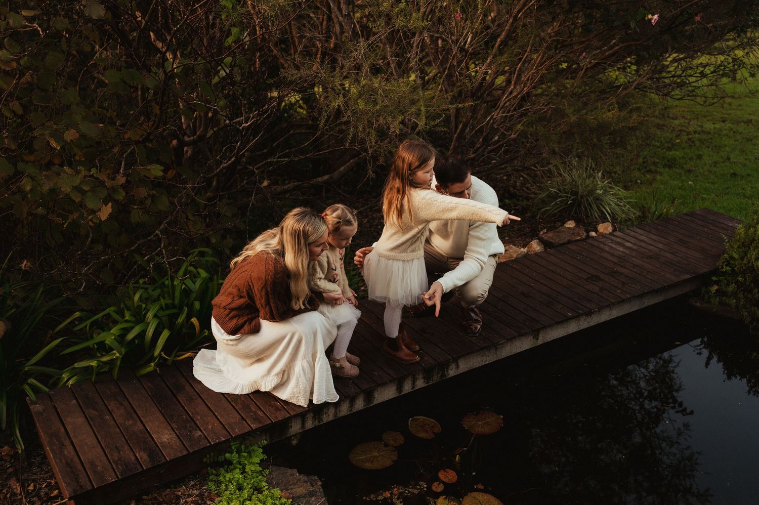 Four people sitting together on a wooden dock bridge surrounded by lush greenery beside a calm pond.