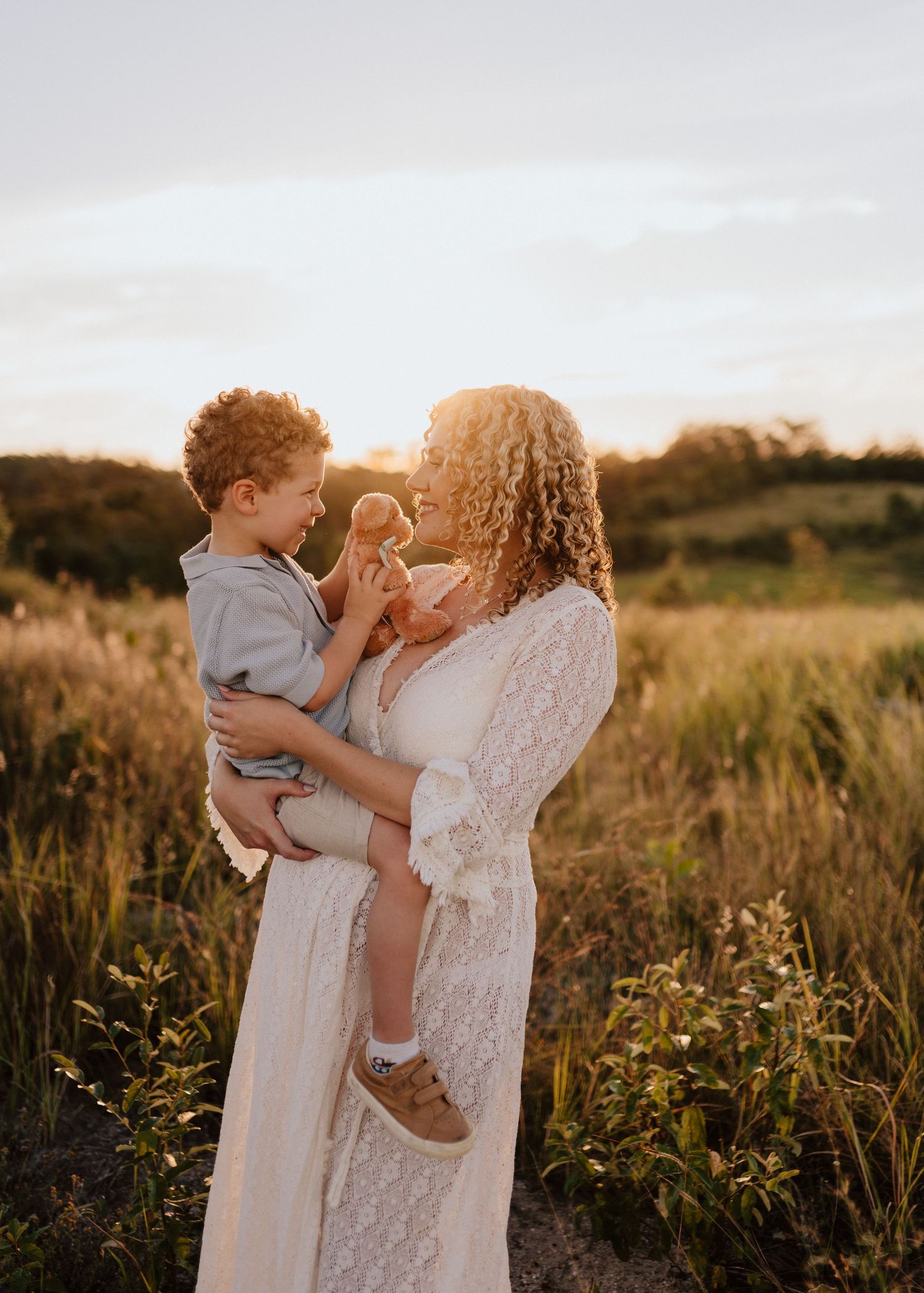 Mother and child share tender moments in a sunlit meadow at golden hour wearing white lace and casual attire.